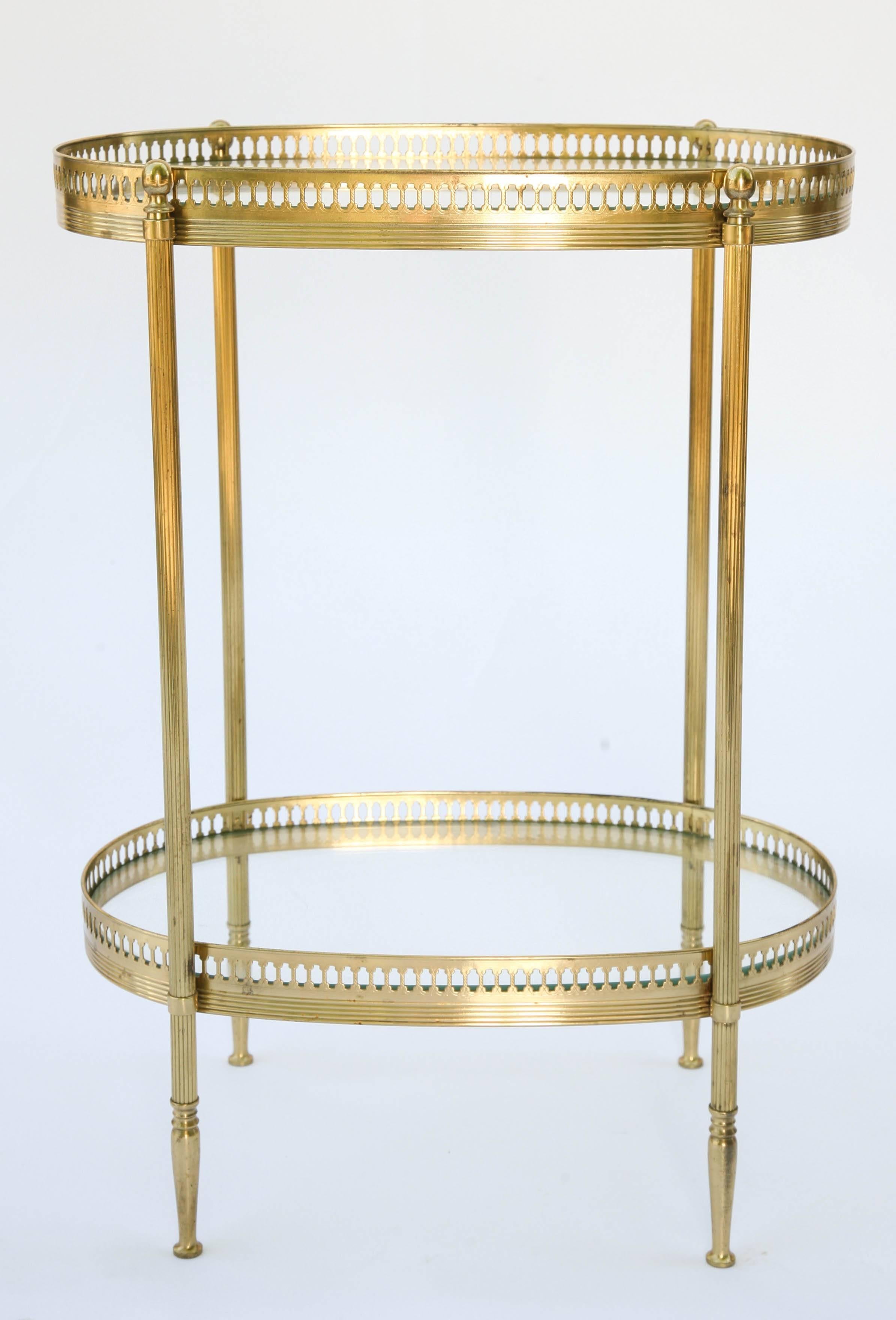 Occasional, or end table, of brass, in the style of Jansen, having two oval tiers, each with pierced galleries and glass tops, raised on round, reeded legs with ball finials, ending in pegged feet.

     