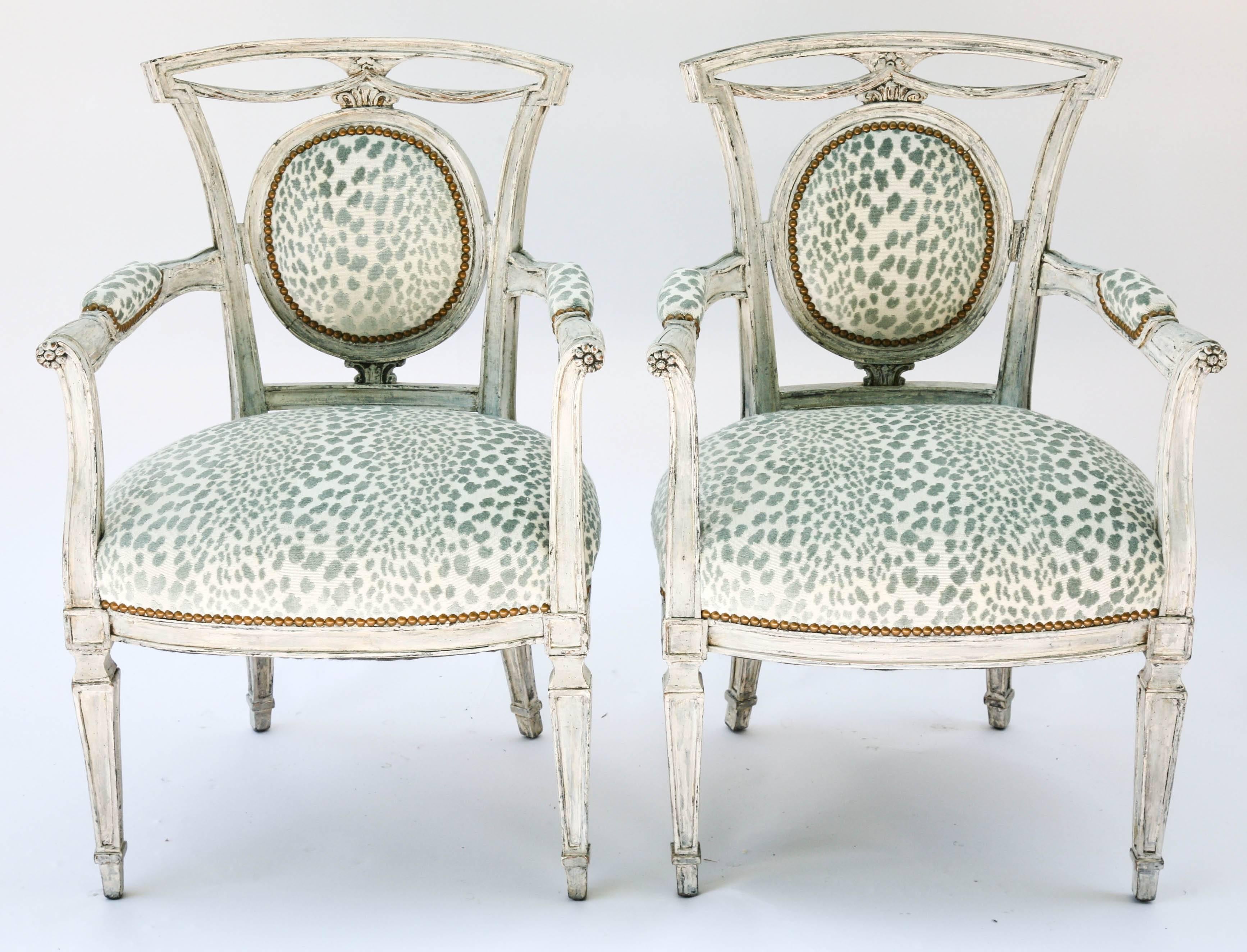 Pair of open armchairs, having a worn-painted finish, each channeled frame inset with round padded back surmounted by carved swags, outswept arms with padded elbow rests, on upturned, scrolling terminals, finished with rosettes, on bowed crown seat,