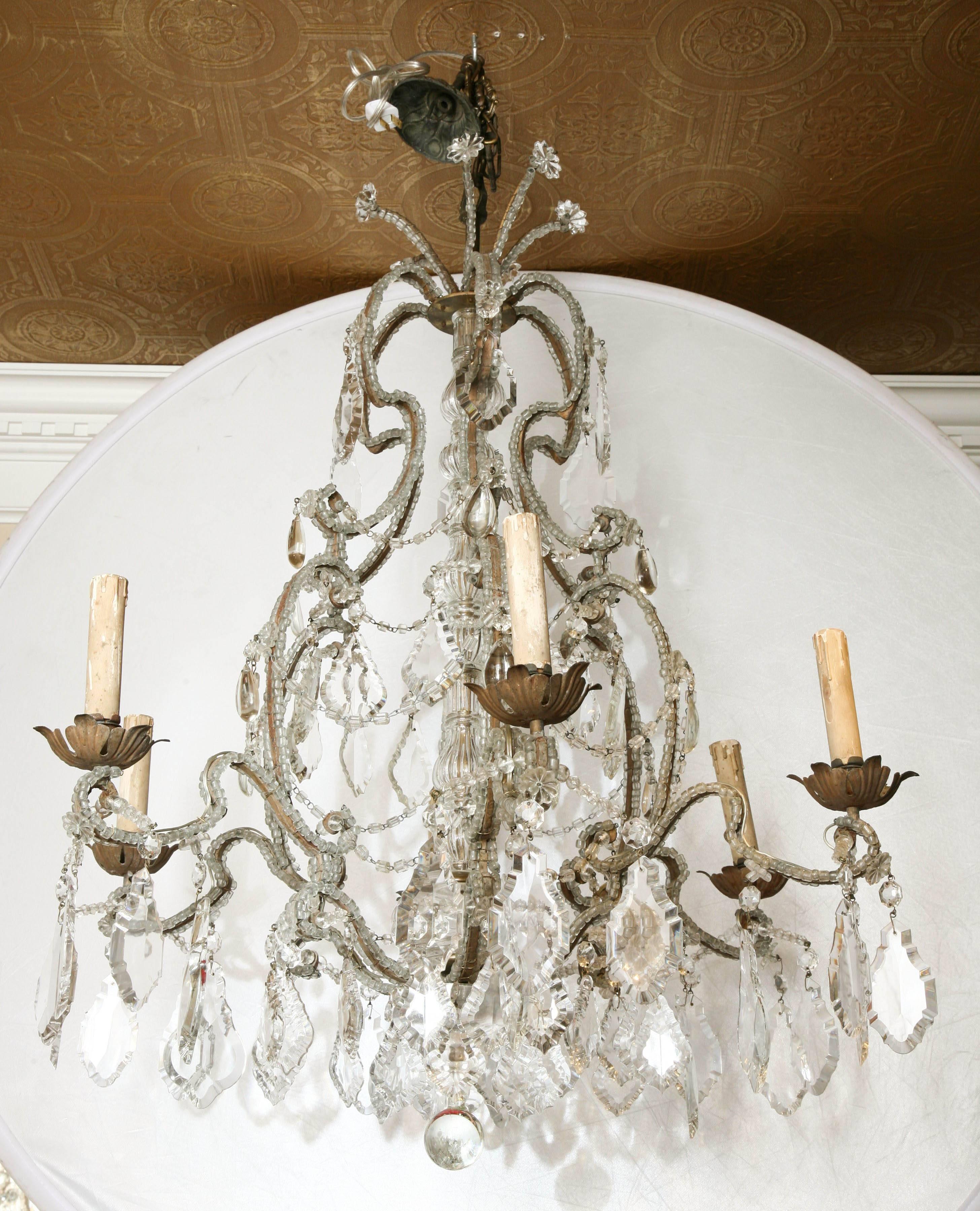 Lampadario con struttura a forma di gabbia, in bronzo, completamente incrostata di perle di vetro macaroni, da cui fuoriescono sei bracci a candela a spirale, che terminano con bobeches foliate, e che circondano la colonna centrale in vetro a