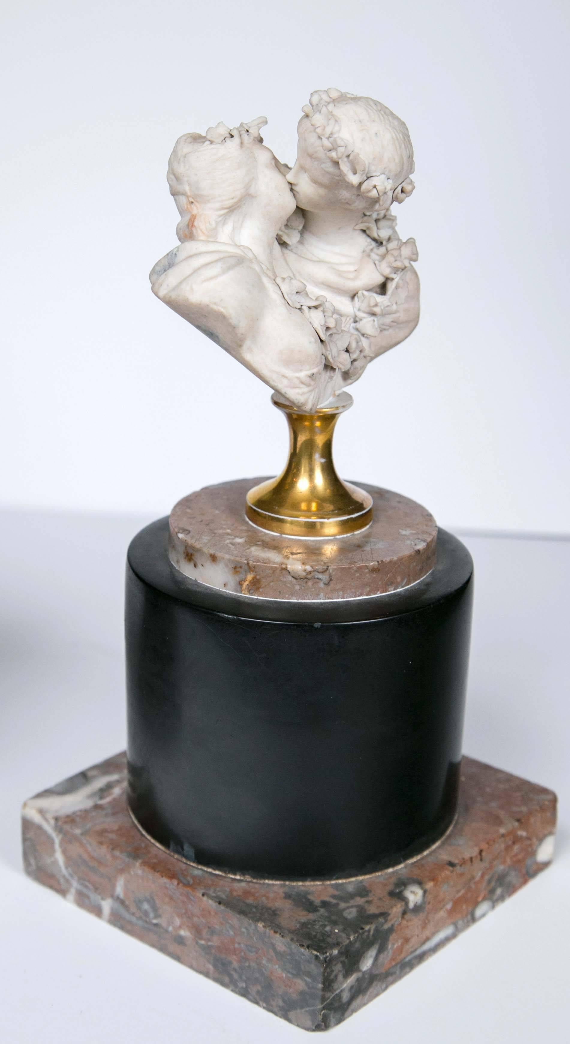 These biscuit porcelain double busts depict young lovers kissing. They are mounted on a brass socle, onto a marble disc which itself is mounted onto a black circular low plinth above a square marble base, Each pair of couples with garlands of
