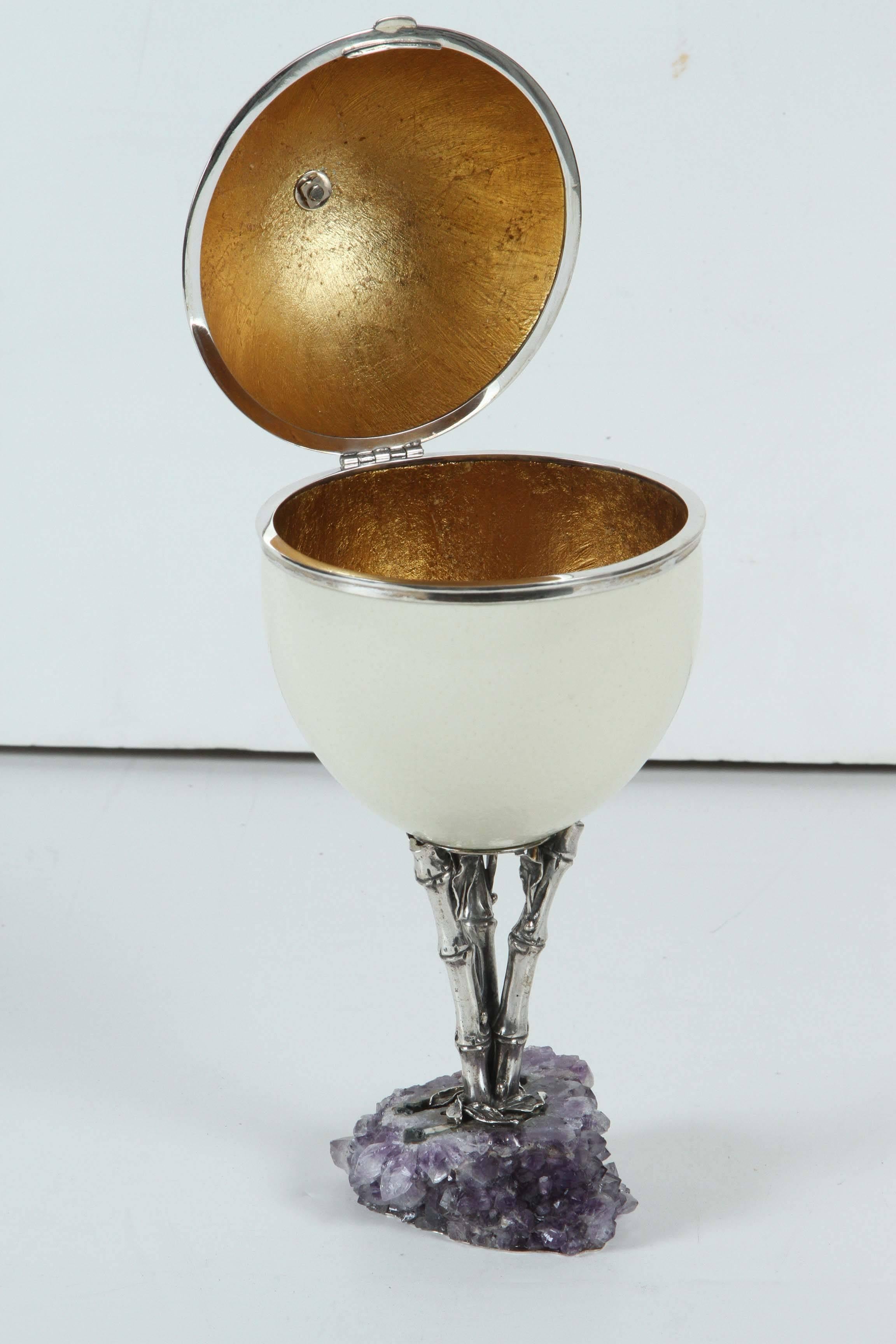 Large, hand decorated ostrich egg container featuring a gilded interior. The whole mounted on a sterling silver pedestal with a multifaceted, amethyst crystal base.