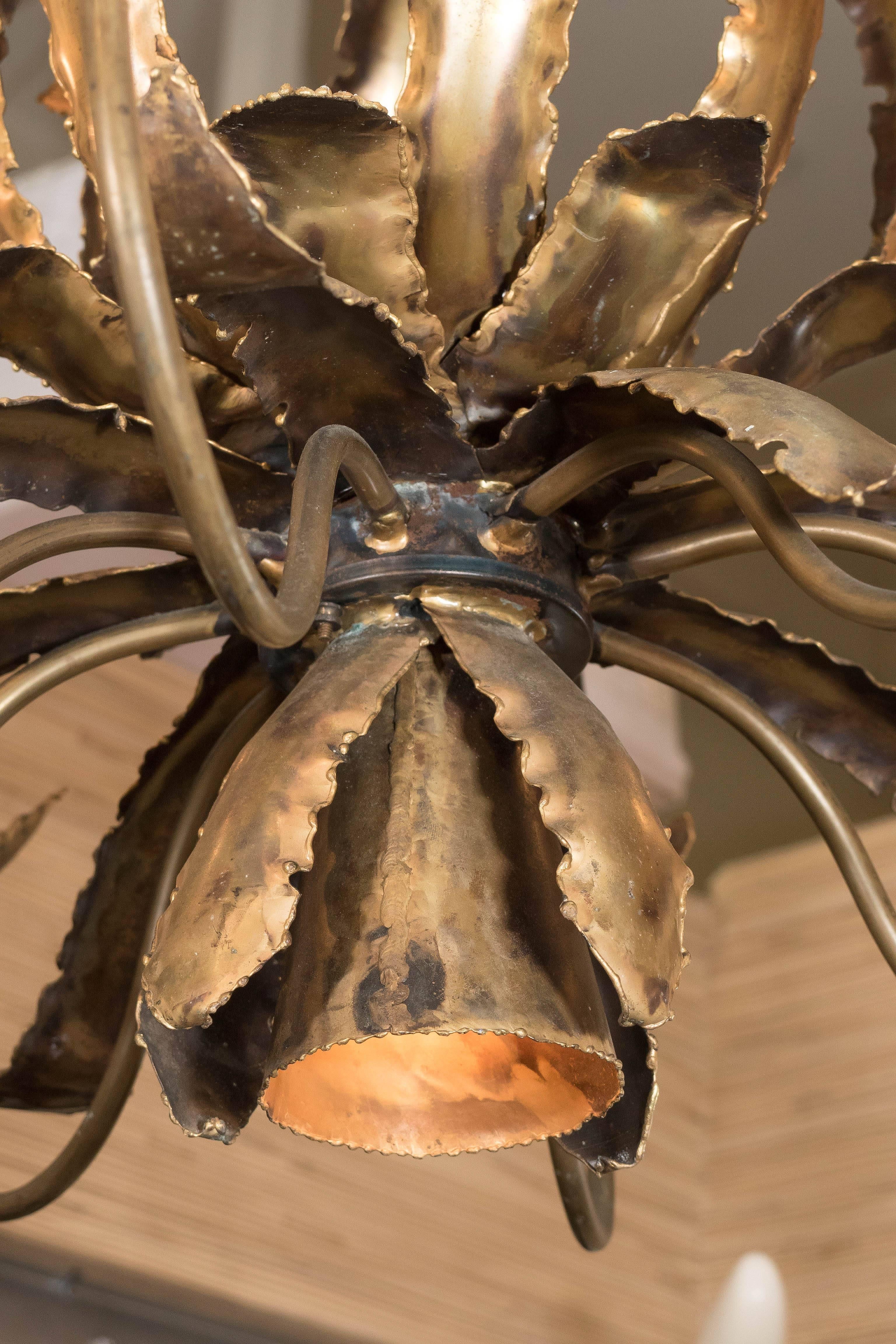 A Brutalist style brass chandelier, with eight scroll arms, each with decorative bobeche and socket cover, surrounding the pineapple form body, composed of torch cut and soldered patinated metal sheets, shaped into curled leaves, giving this light