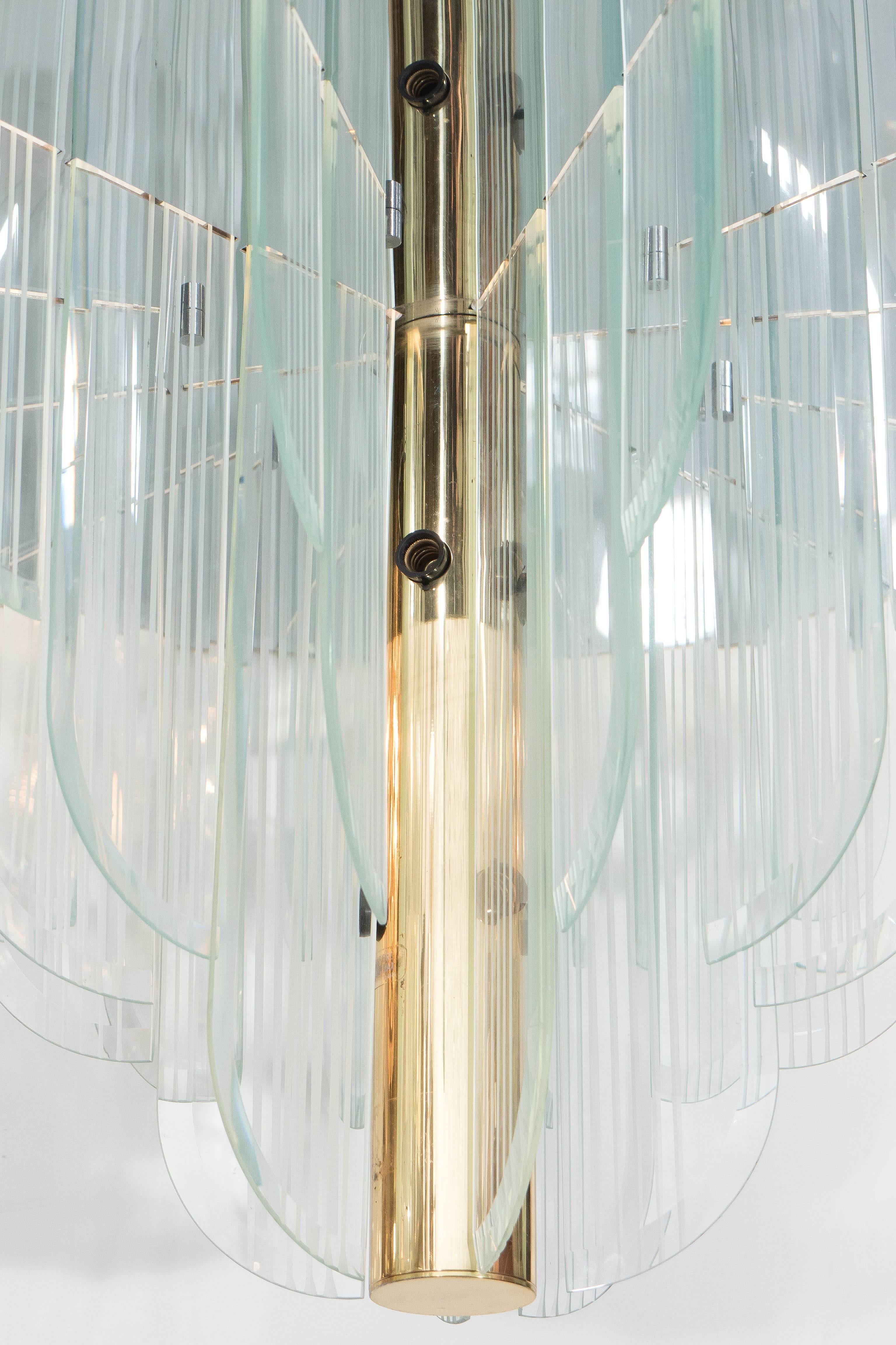 An Art Deco style chandelier, produced circa 1970s by Lightolier, featuring curved glass panels, affixed to a round canopy with reflective plate, supported by a cylindrical nucleus in brass with surrounding sockets. This light fixture remains in