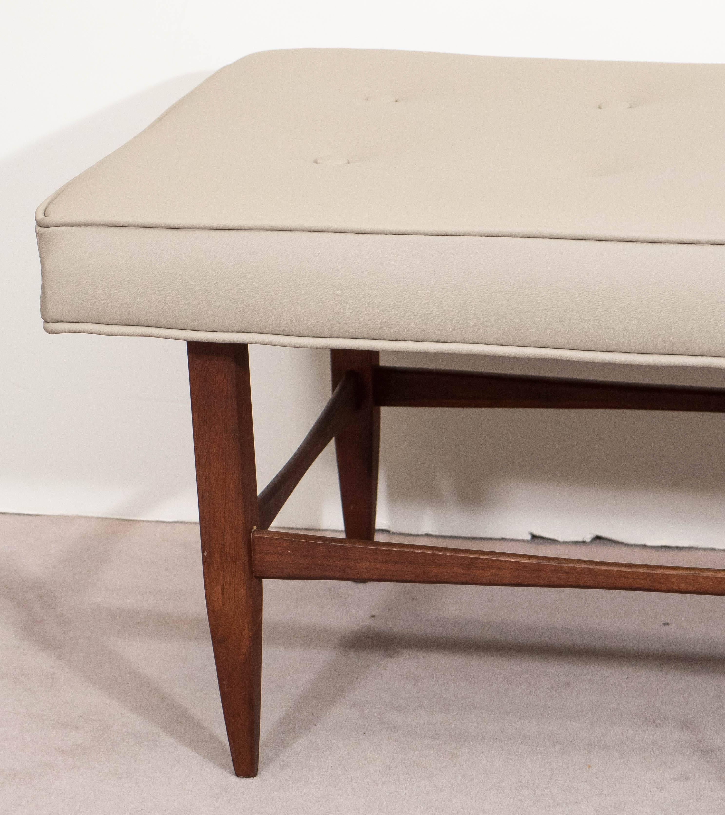 A vintage lengthy bench, with cushioned tufted seat in beige vinyl, against a wooden frame, on six squared tapered legs, with curved stretcher bars. This bench remains in excellent condition, with some presence of wear to base consistent with age