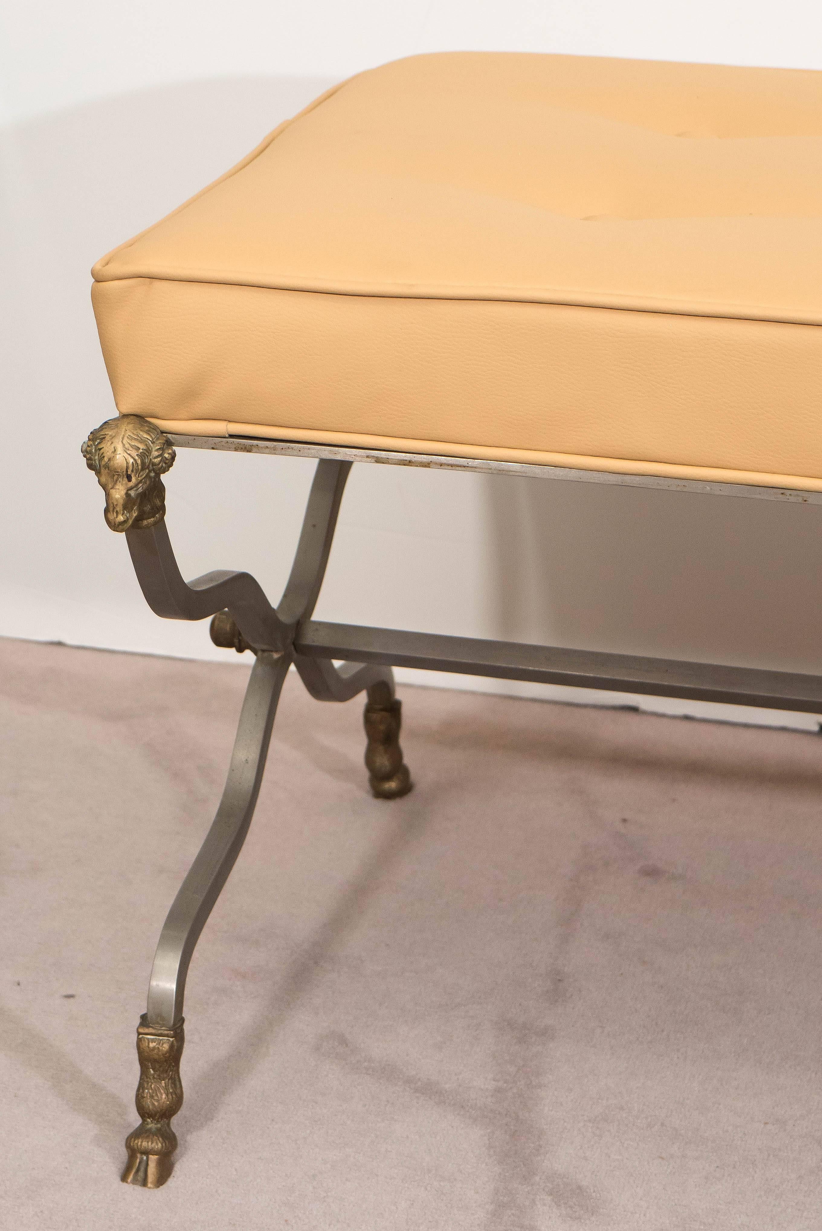 This Hollywood Regency style bench, produced in Italy circa 1950s-1960s, includes tufted cushion seat in light yellow vinyl, on brushed steel frame with three curvaceous X-form legs, each detailed with ram's head and terminating in hooves in brass.