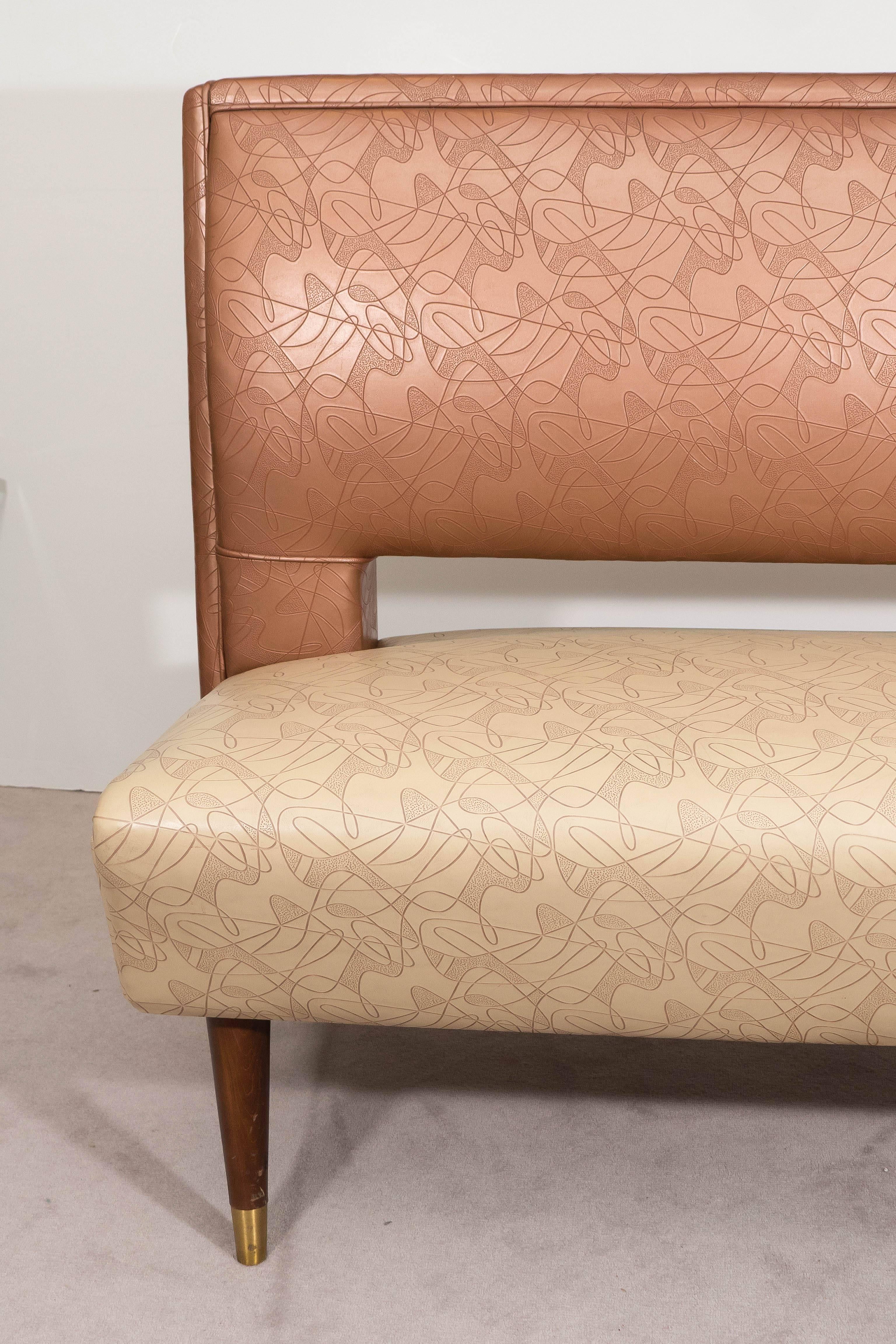 A settee or banquette circa 1950s, manufactured by the Brody Seating Company of Chicago, Illinois, upholstered in vinyl, the back and seat in tones of copper and beige, impressed with Googie-style patterns, on tapered wood legs with brass sabots.