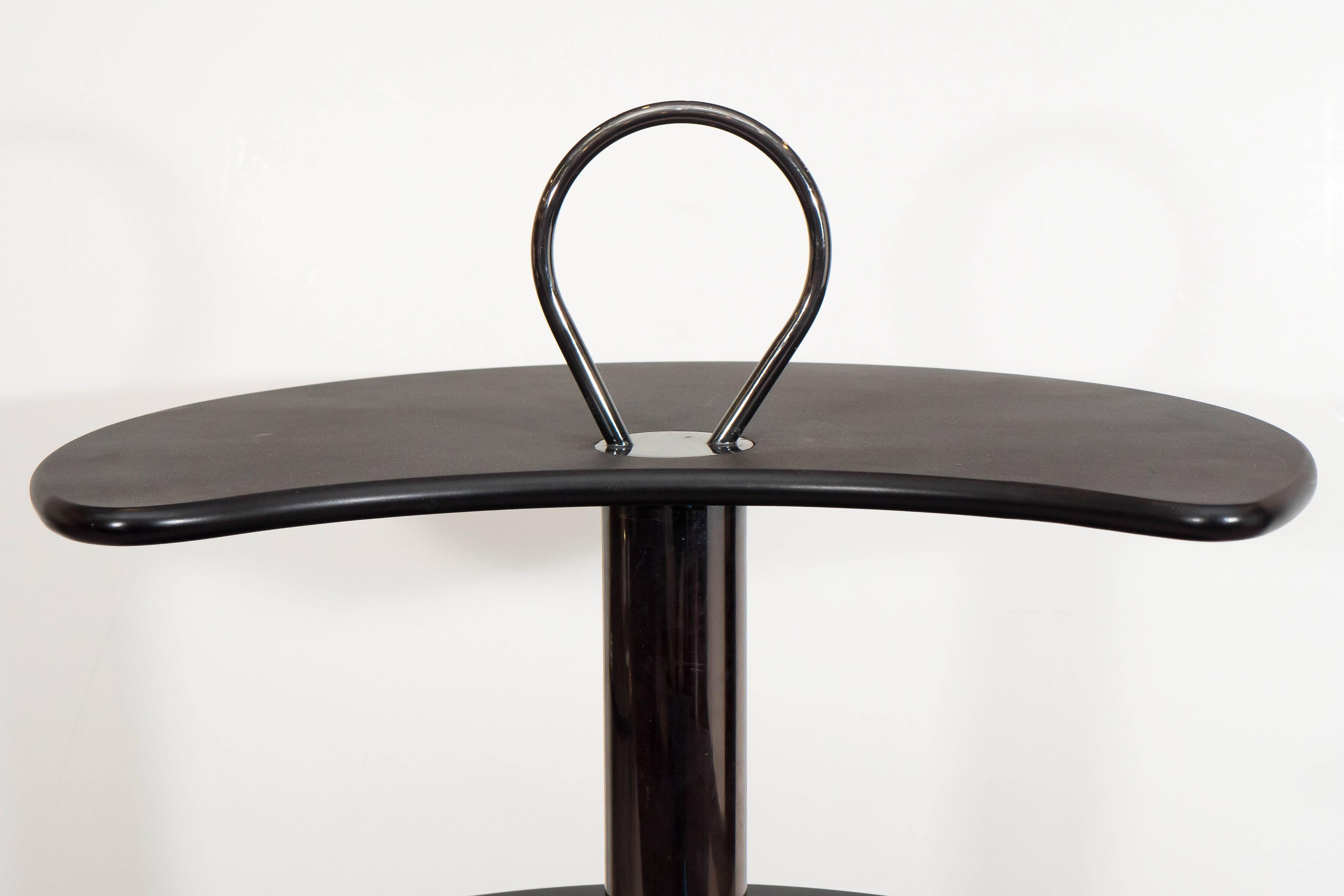 This circular bar cart by the Design Institute of America (DIA), comes in black lacquered wood, with three-tiers of revolving shelves, surrounding a central pole with round handle, on casters. Markings include label [Design Institute of America