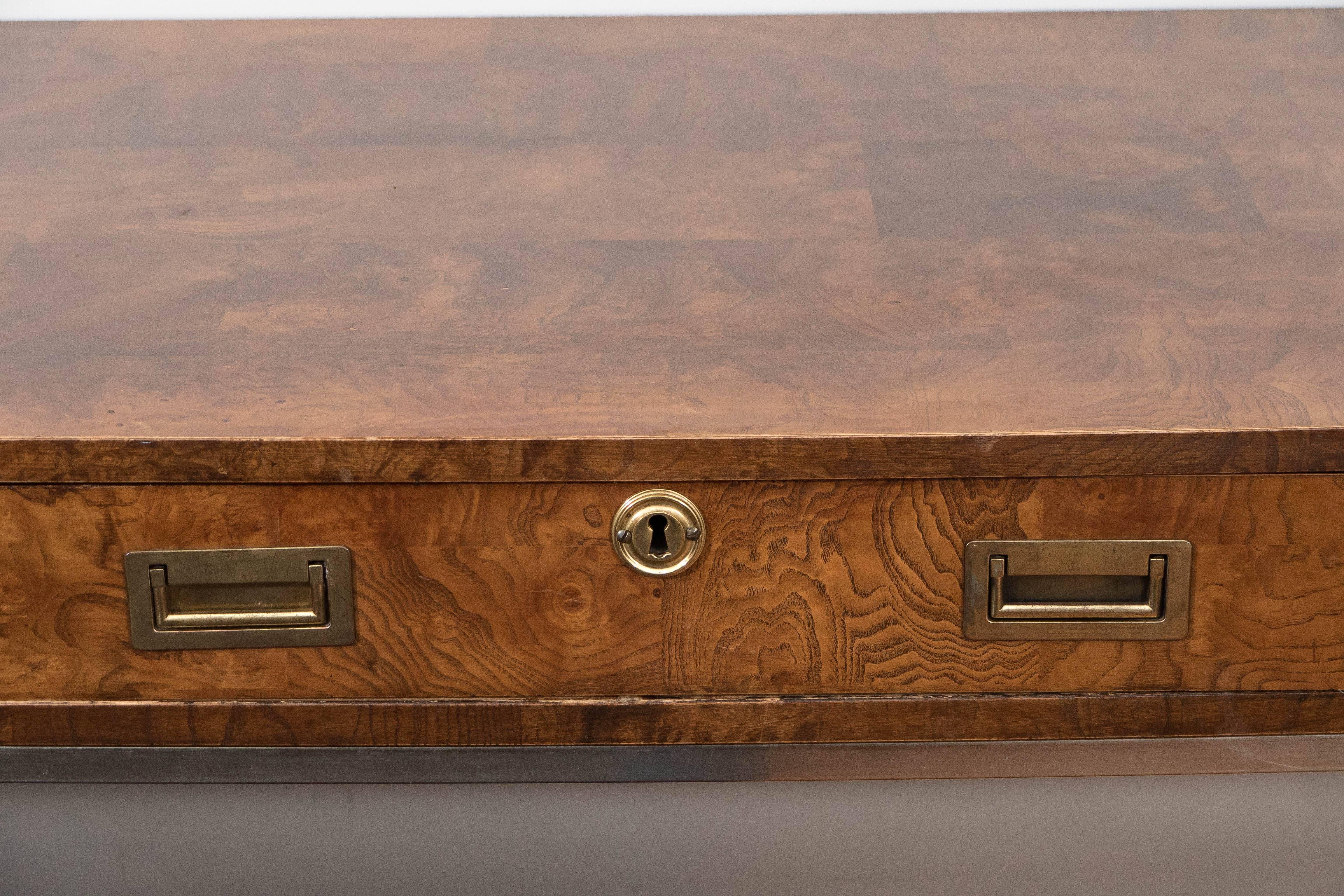 A campaign style desk, produced circa 1960s by Bernhardt furniture company, veneered in panels of highly detailed wood with brass accents, including three drawers with recessed handles, escutcheon to center, on highly linear metal base. Markings