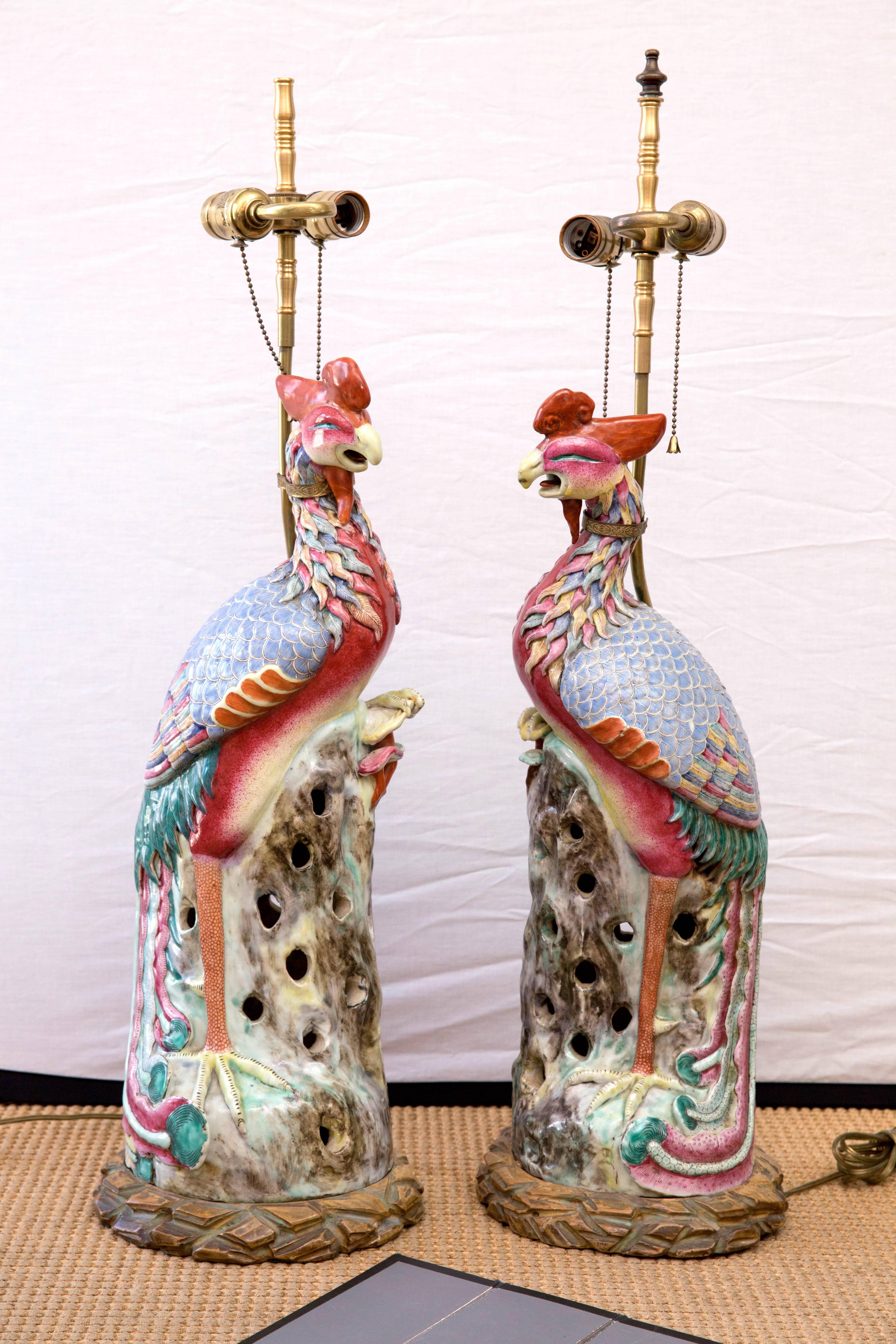 Chinese porcelain figurines of phoenix birds standing on a rocky outcrop. The pair are mounted on carved wood bases and mounted to brass for use as double socket table lamps. Polychrome decorated in famille rose colors. The birds themselves are 24