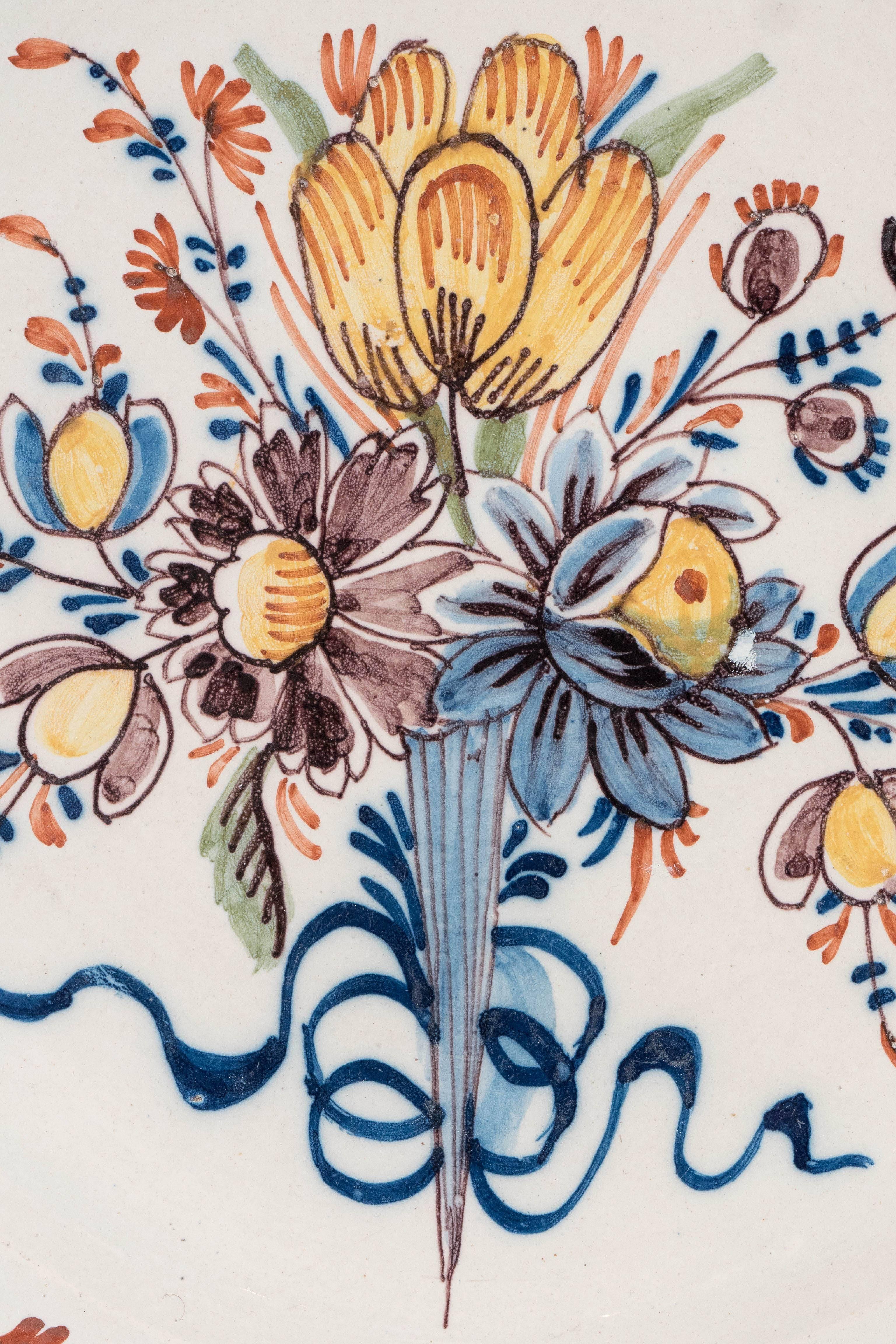 A late 18th or early 19th century Dutch delft charger featuring a bouquet of brightly colored tulips and other flowers painted in yellow, manganese, and blue. The border is painted with flower heads and scrolling vines in the same colors.