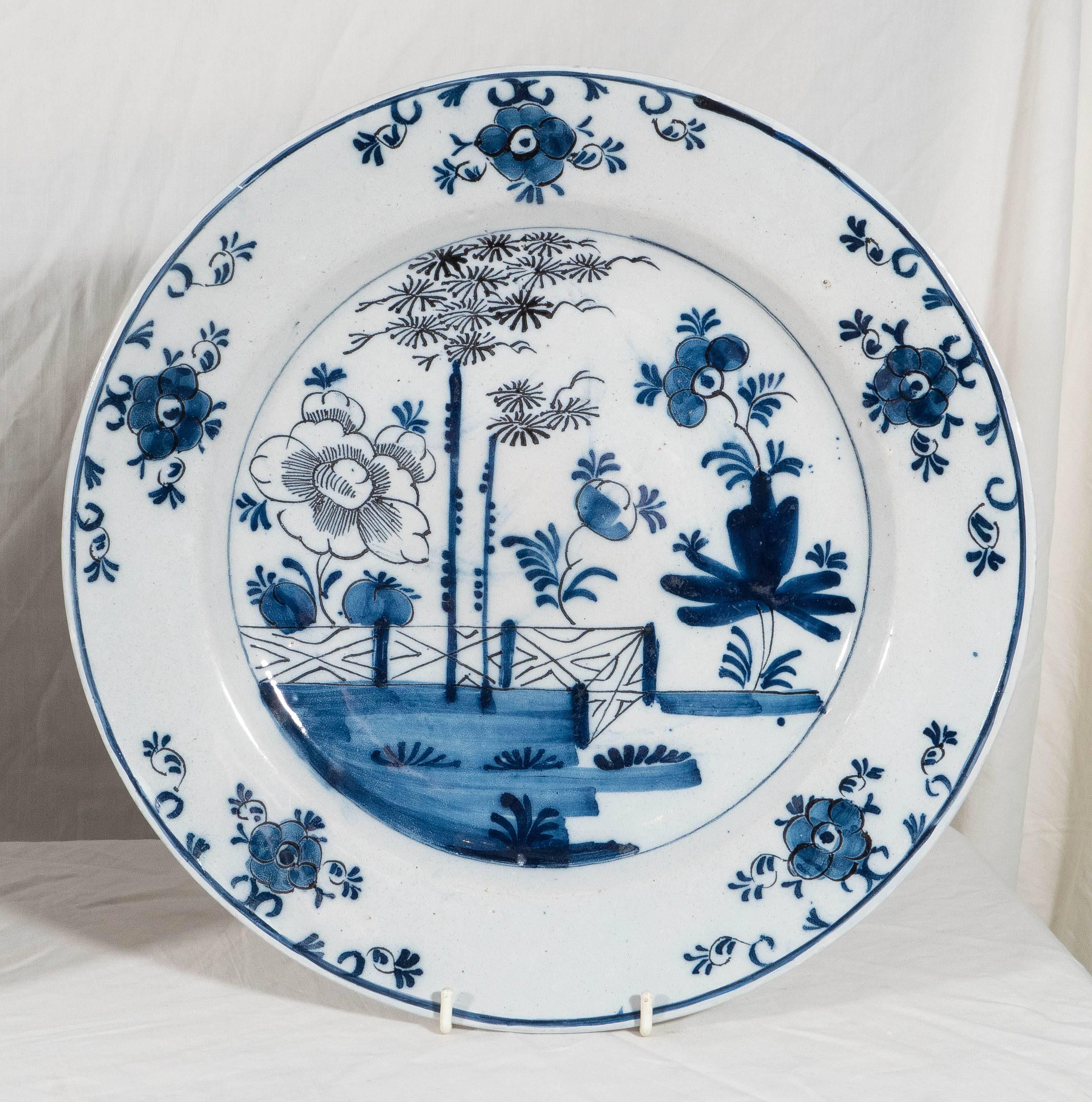An 18th century pair of blue and white Delft chargers hand-painted with a chinoiserie garden scene. The scenes show an oversized peony, a garden fence and at the center two tall trees. The borders are painted with five flower heads and scrolling