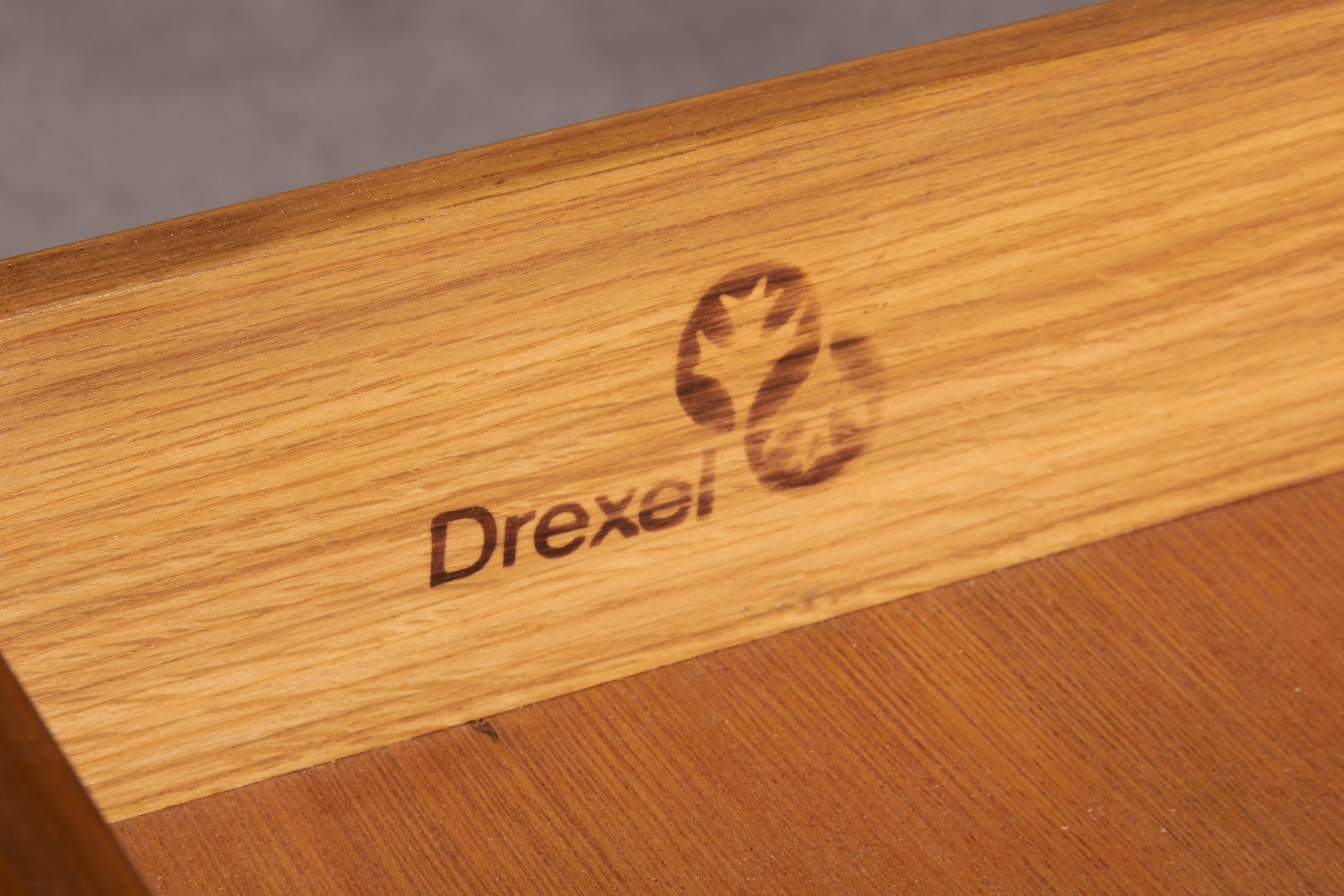 This 1970s nightstand by Drexel comes in warm toned wood, with artful bookmatched veneer top on rectilinear legs and crossed stretcher; single drawer with brass pull included. Markings include [Drexel] hallmark stamped to drawer interior,