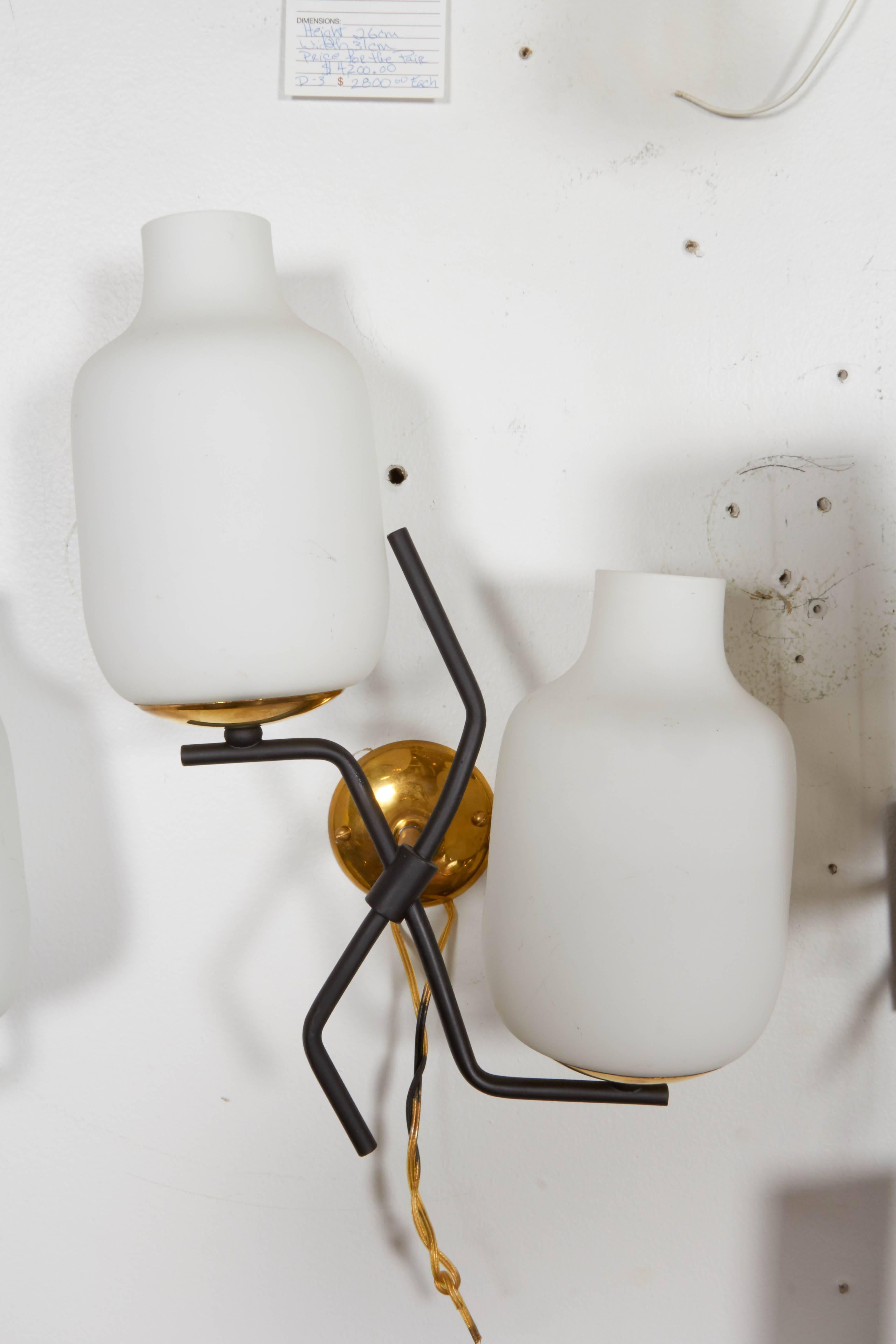 A pair of, circa 1950s Italian wall sconces by Stilnovo each with two milk glass shades, on contrasting black enamel stems, branched out at dissimilar lengths from brass mounts, creating a great sense of proportion. Wiring and sockets to US