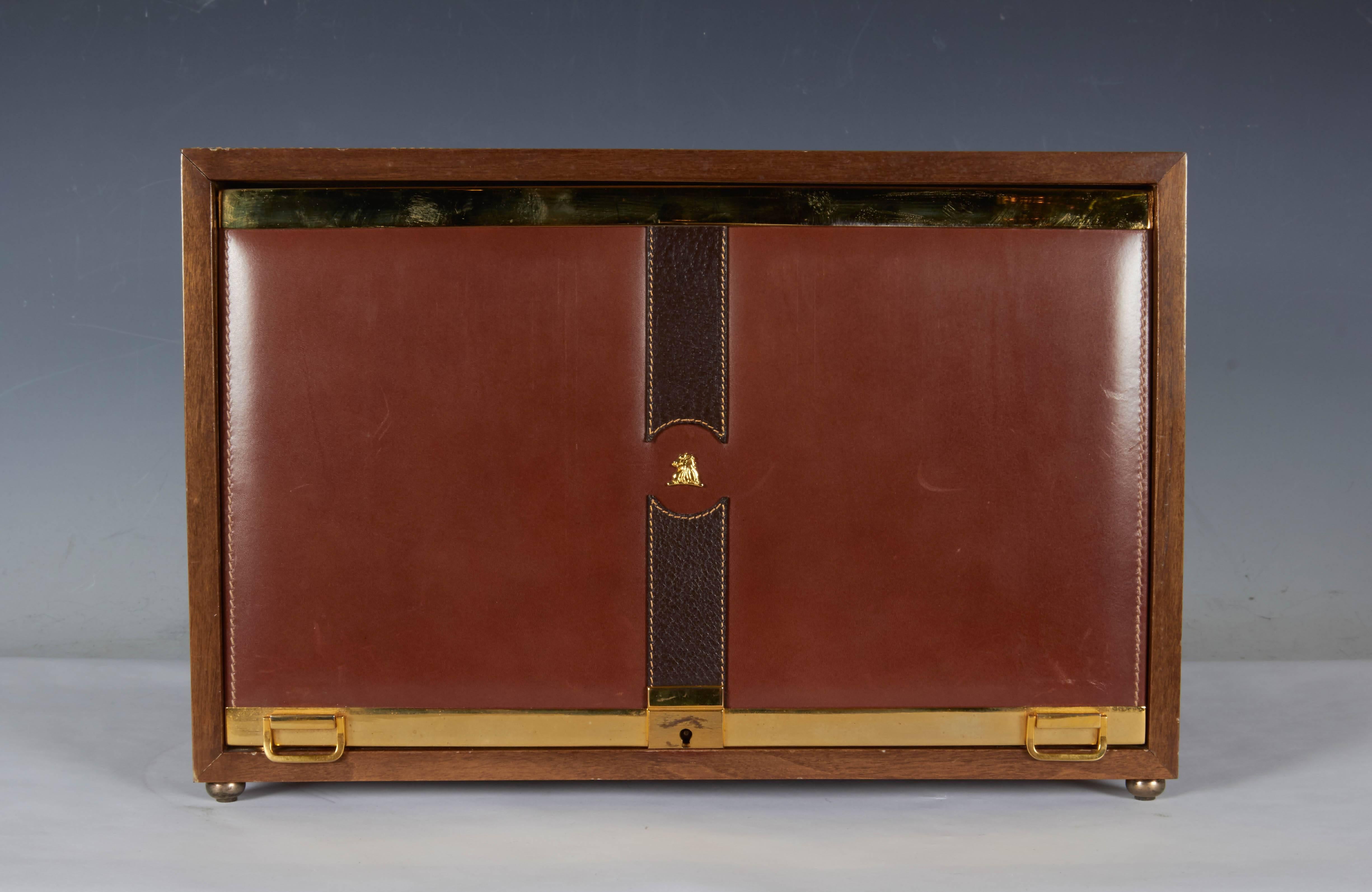 This circa 1980s Italian men's jewelry box by Mark Cross, comes in leather, in Dual tones and textures of warm and dark brown against a wooden frame, brass hardware and handles to the front and sides, on ball feet; included is a locking panel, which