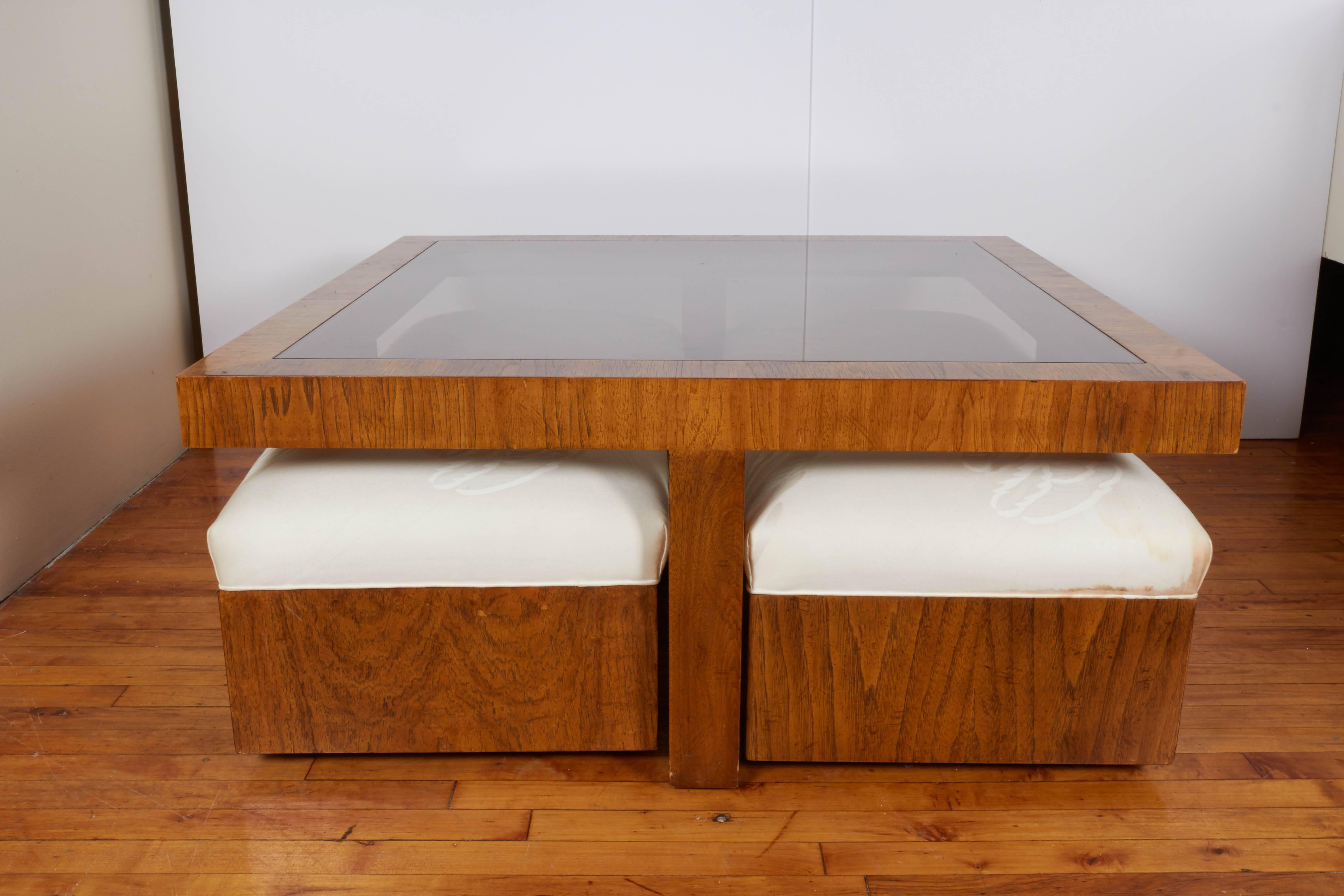A Drexel cocktail table from the Consensus collection, produced, circa 1970s-1980s, with smoked glass top, against a beautifully veneered wood frame with crossed base, providing space for the included set of four ottomans, each with upholstered