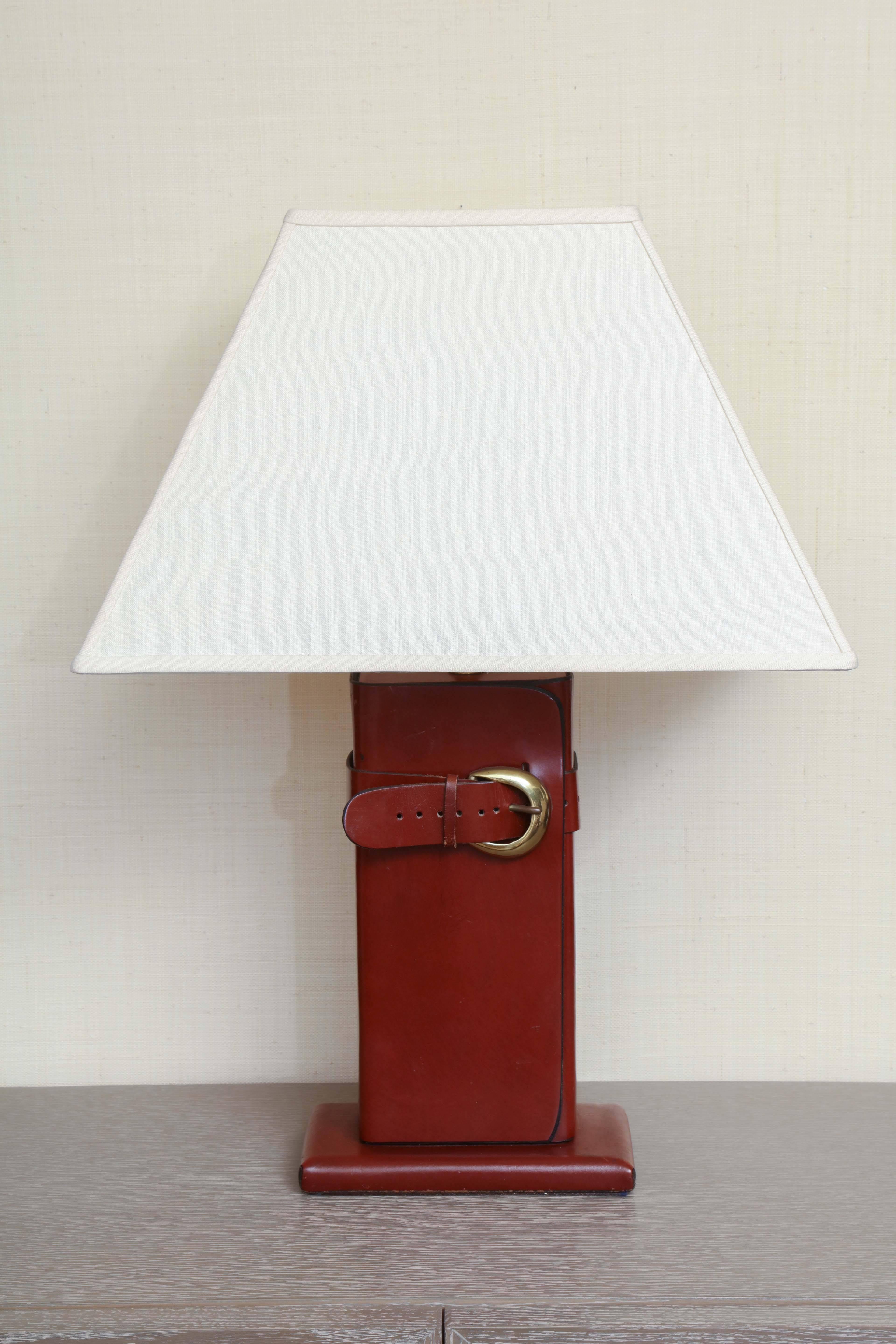 Leather lamp with brass buckle and ivory linen shade in the style of Hermes, circa 1960.