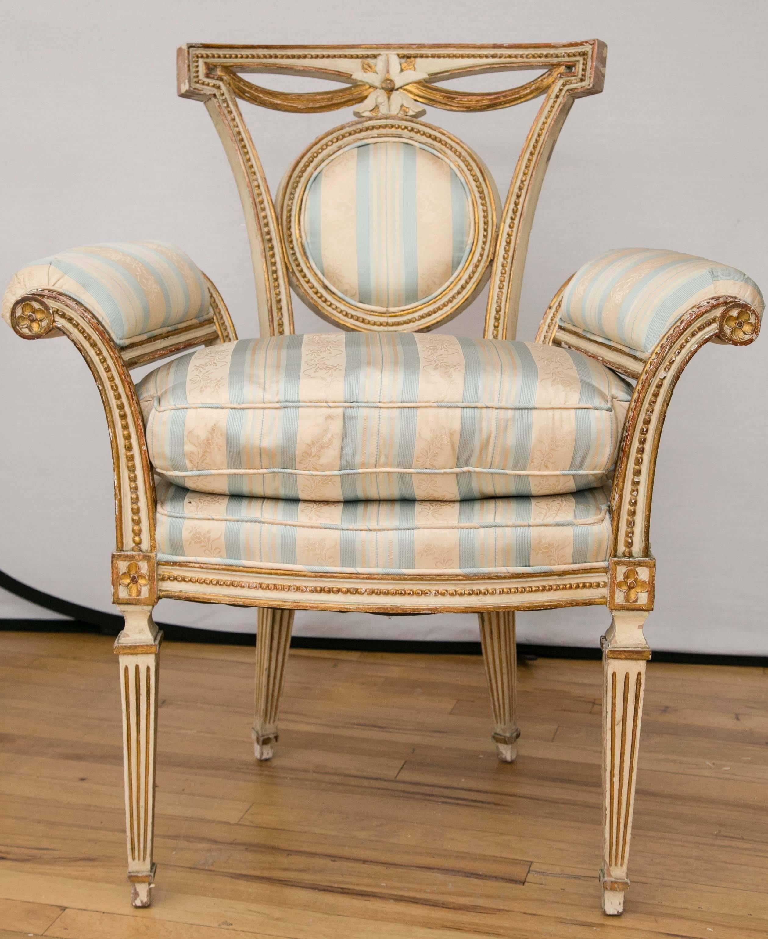 A pair of painted and gilded wood medallion back chairs. Delicate bead and swag detail surround a central flower. Padded scroll form arms sit atop tapering fluted legs.