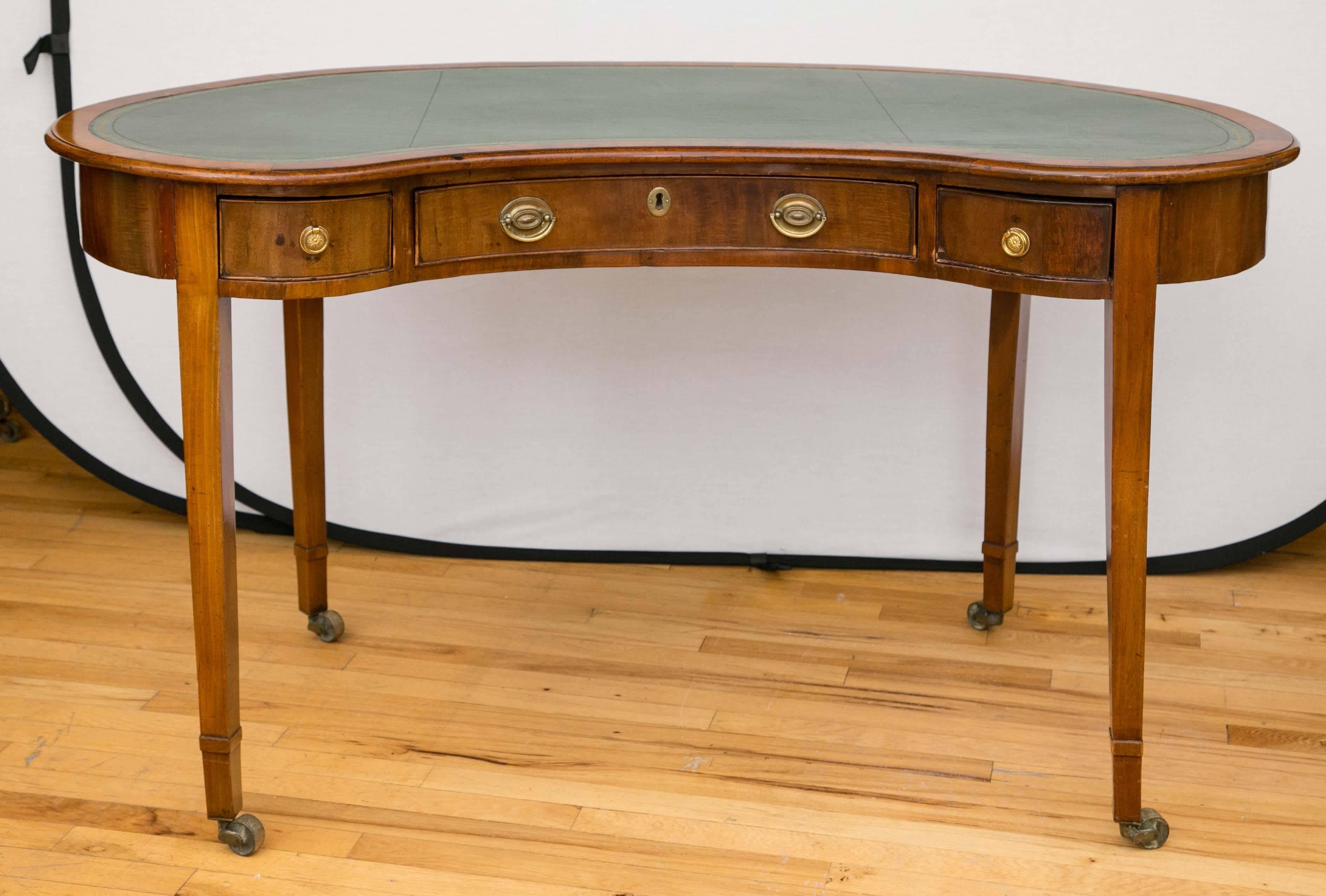 A 1790s English rosewood writing table in the Hepplewhite style. The apron is fitted with a large center drawer and two smaller drawers on either side. The table stands on tapering legs that terminate in brass castors. The inset tooled and gilt