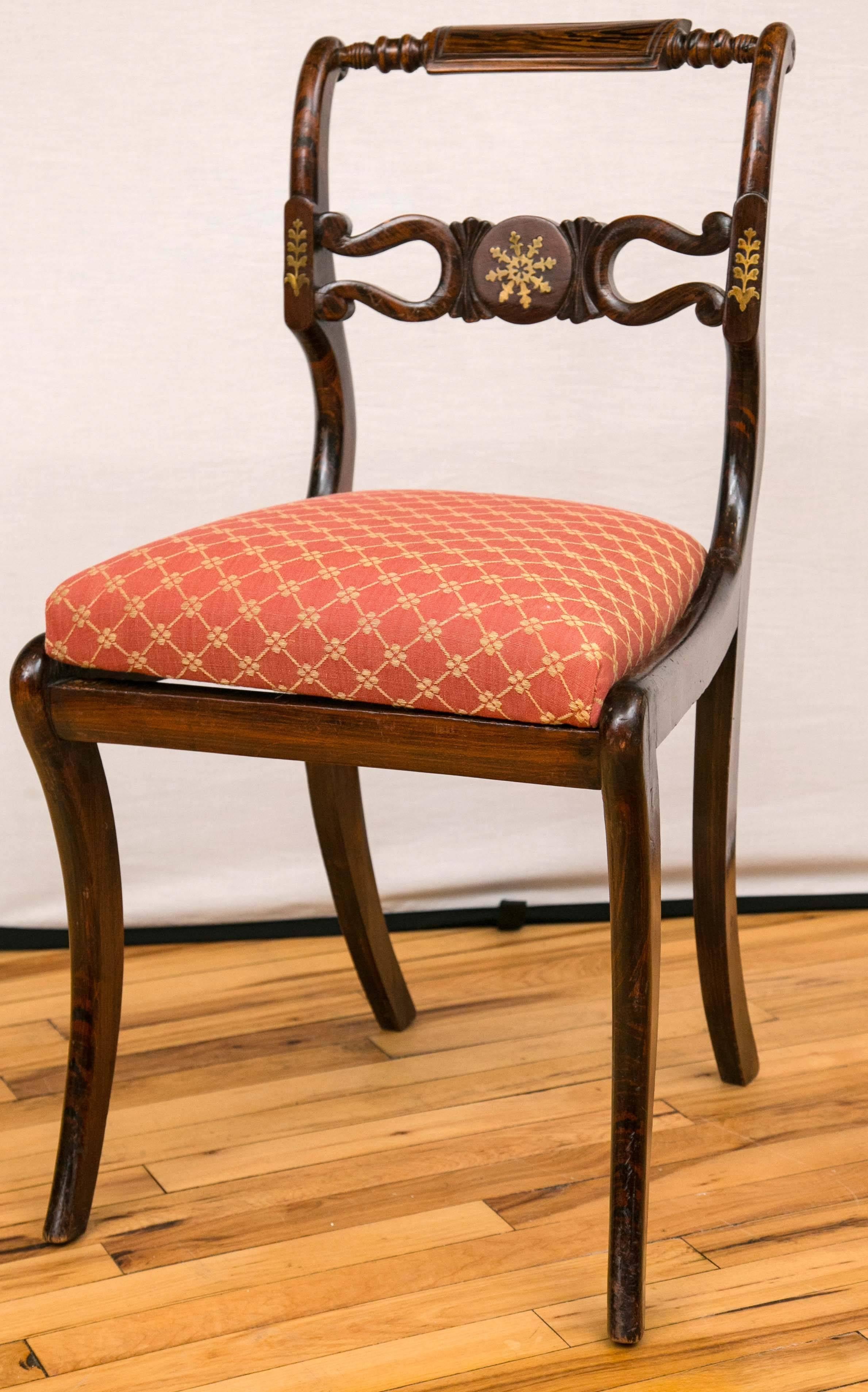 A set of four Regency period side chairs, with turned crest rails. The central splats also with brass inlay. Drop in upholstered seats over saber form legs.