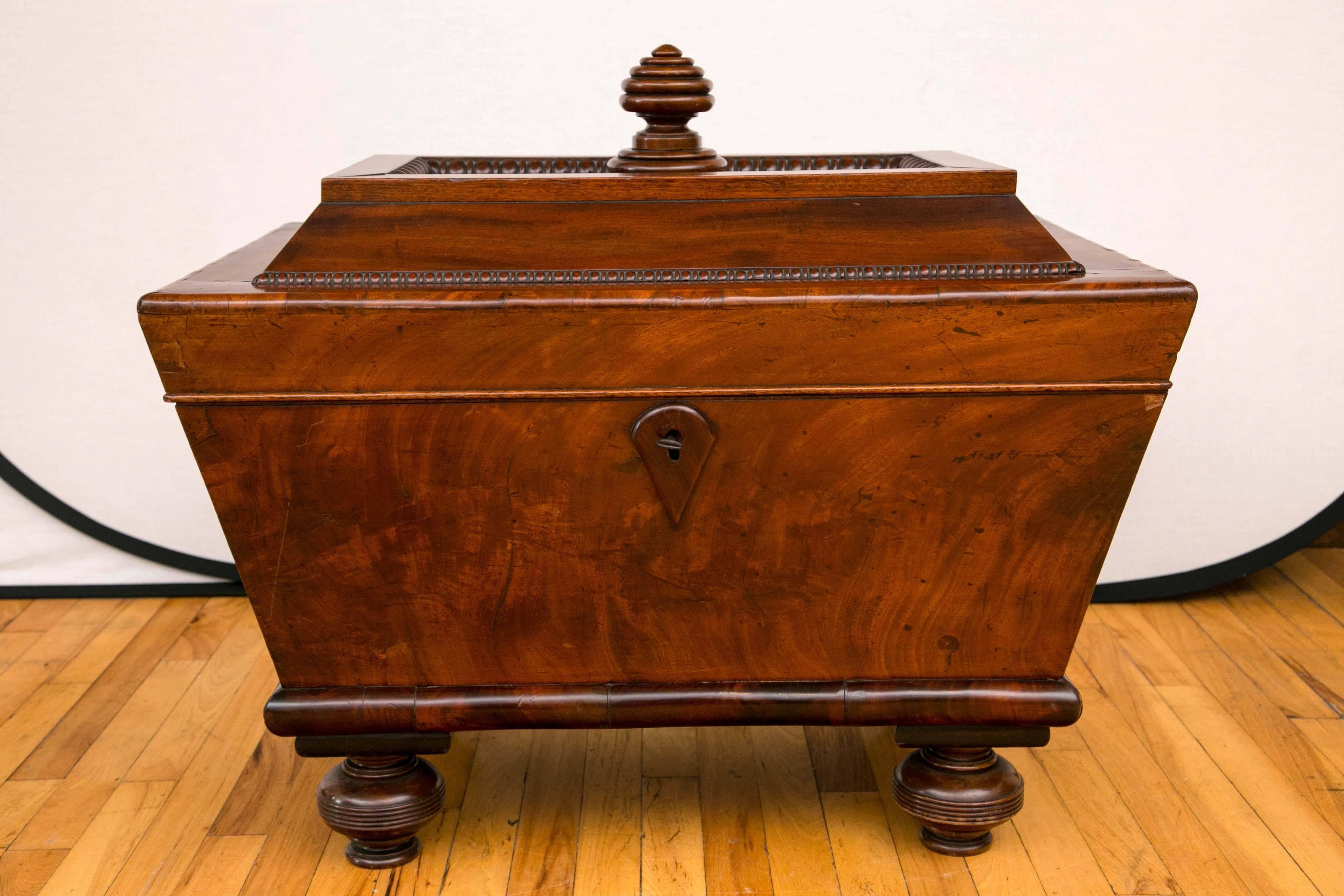 An early 19th century mahogany wine cooler of sarcophagus form, the raised hinged top above the body with recessed panels on a platform base with turned ball feet. The raised top surface is defined by a turned "honeycomb" finial. Each side
