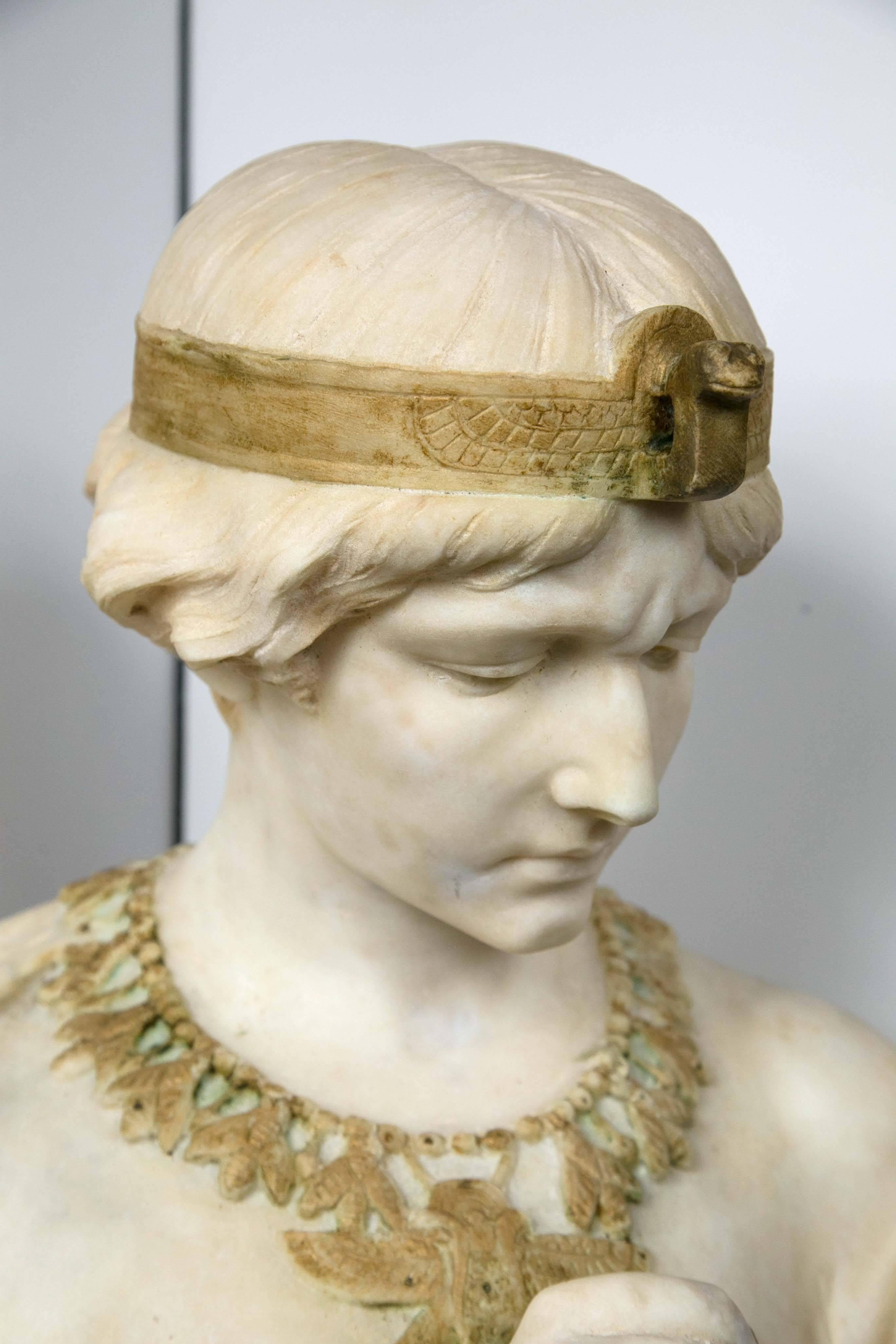 Beautifully carved unusual bust with contrasting marble highlights of an arm band with scarab, head band with snake head, necklace of bees (?) and drop central medallion with wings, and the asp in her right hand. Her hair tied in a bun. Wearing a