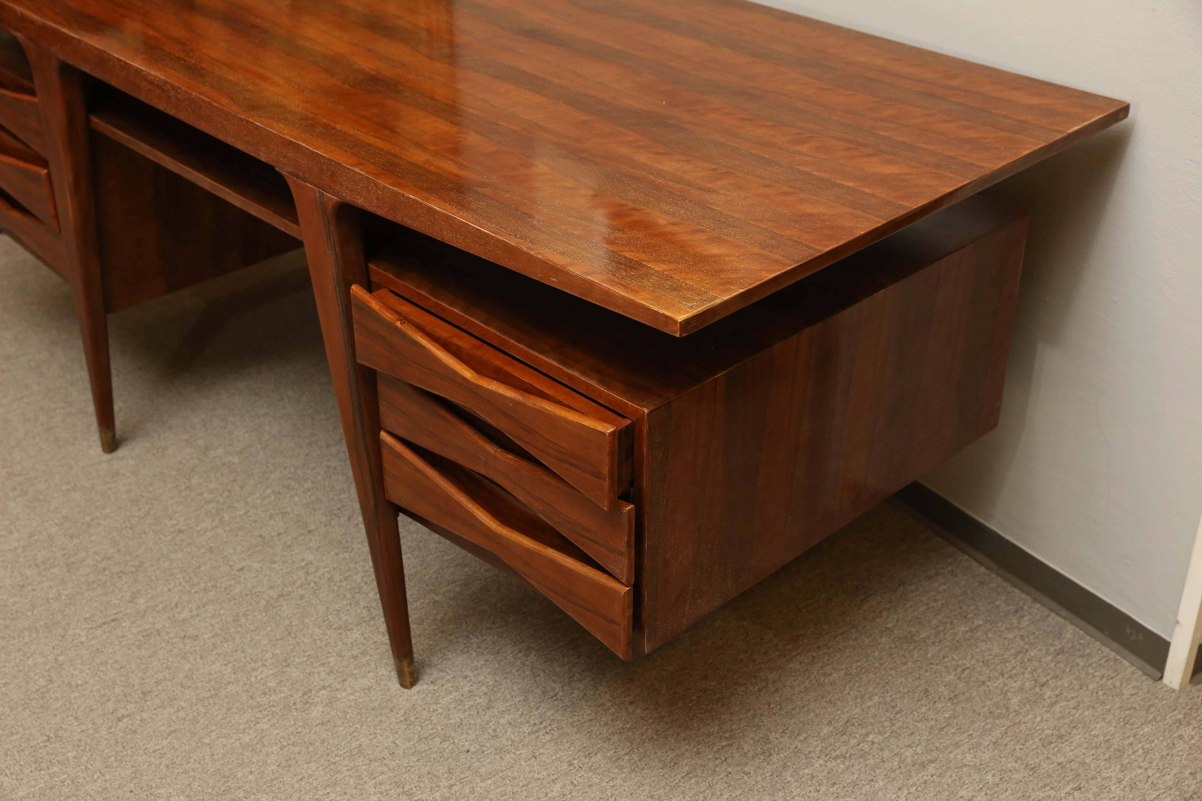 The wide tabletop is presenting a fine walnut wood. Right beneath the top there is wide shelf. Three drawers are attached on either side. Each of the drawers has “bow” shaped handle.
Four slim elongated legs are supporting the desk. On the bottom