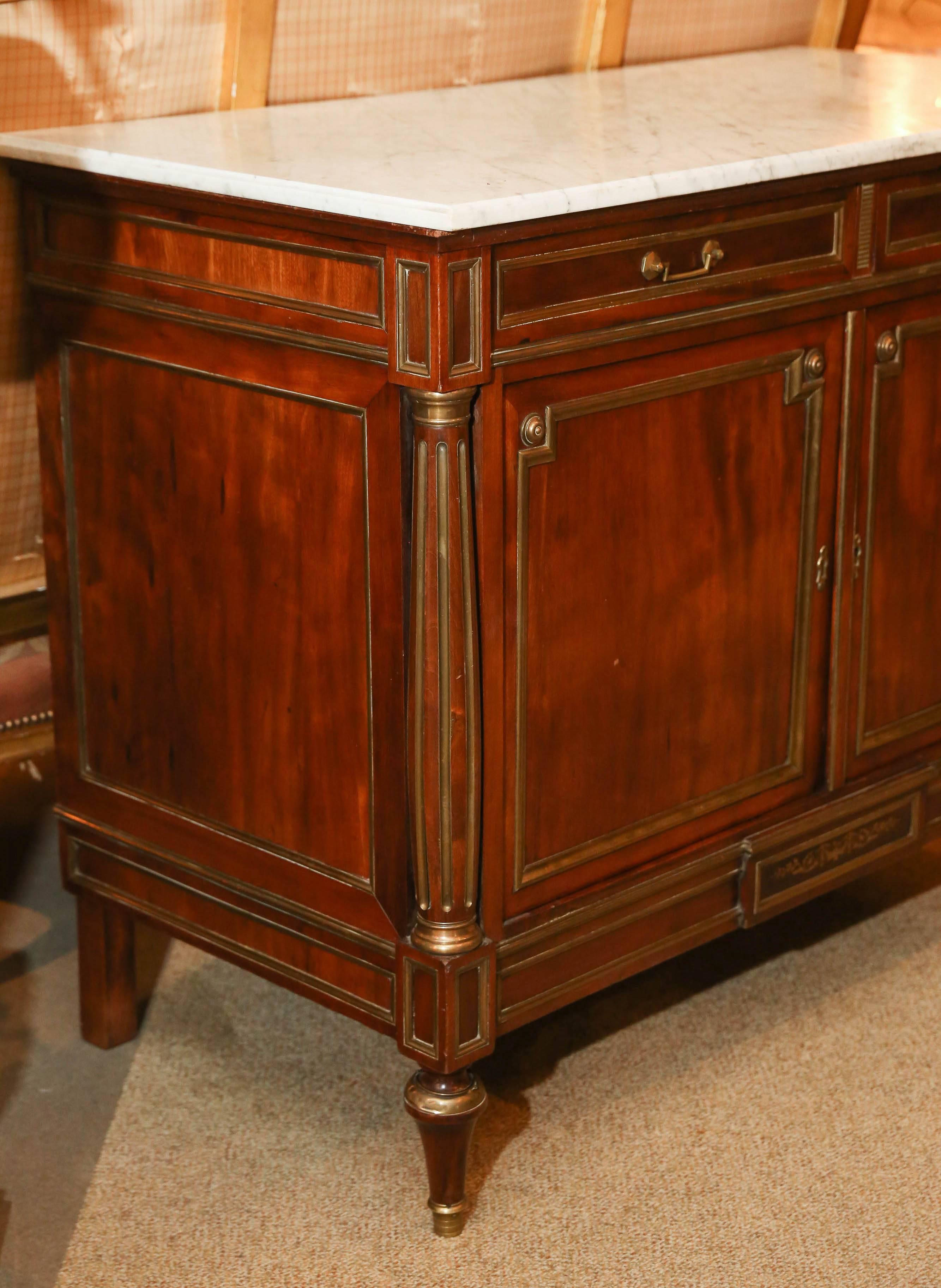 Early 20th century, white marble top with a molded edge, above a case
fitted with two drawers over two paneled cabinet doors, all with brass banding
flanked to either side by a flute upright, raised on brass accented toupee feet.