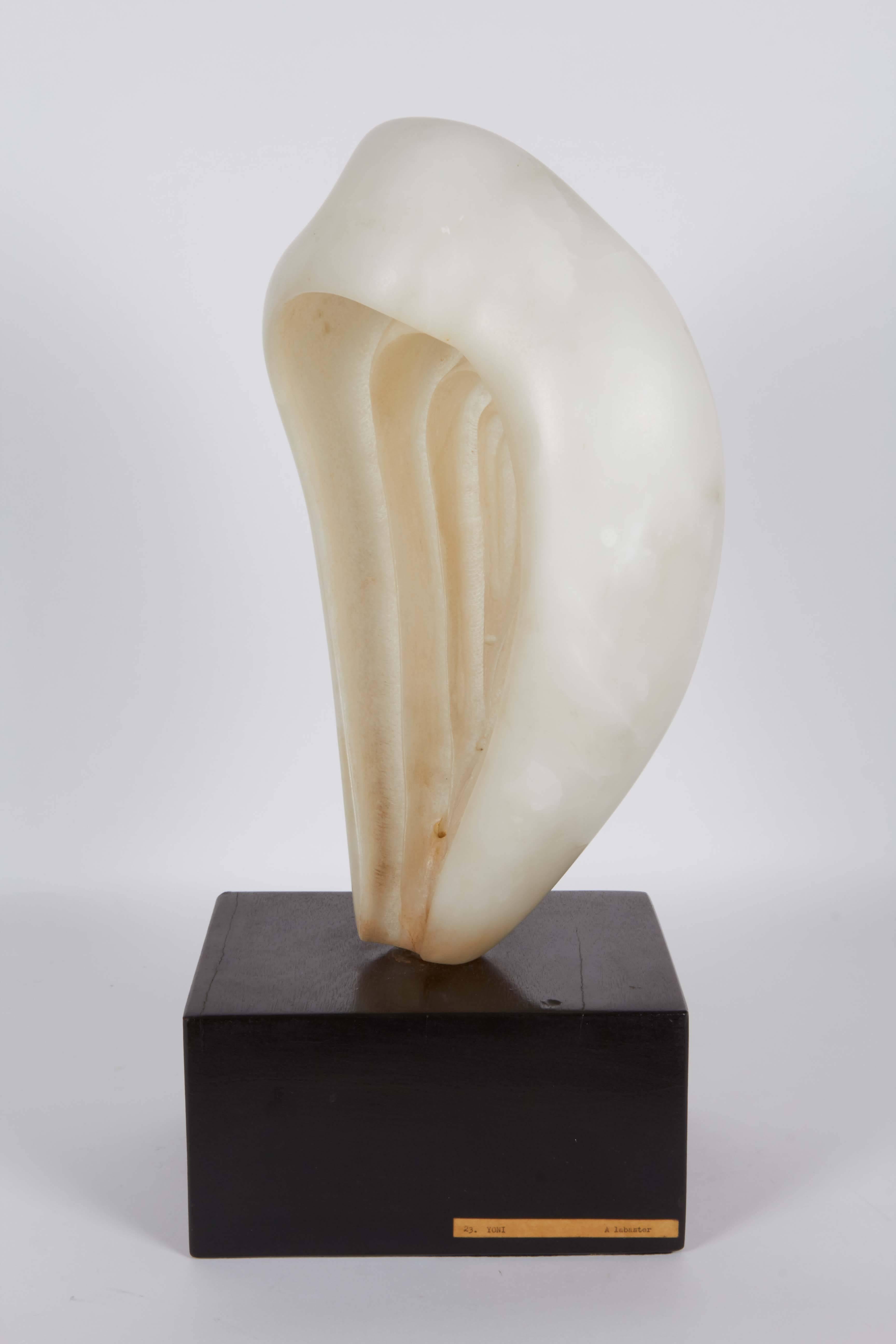 A modern abstract sculpture by an unknown artist, depicting a stylistic yoni symbol, crafted of alabaster, mounted on a rich toned wood base. Markings include a label reading [123. Yoni Alabaster] affixed to the base. Very good overall condition,
