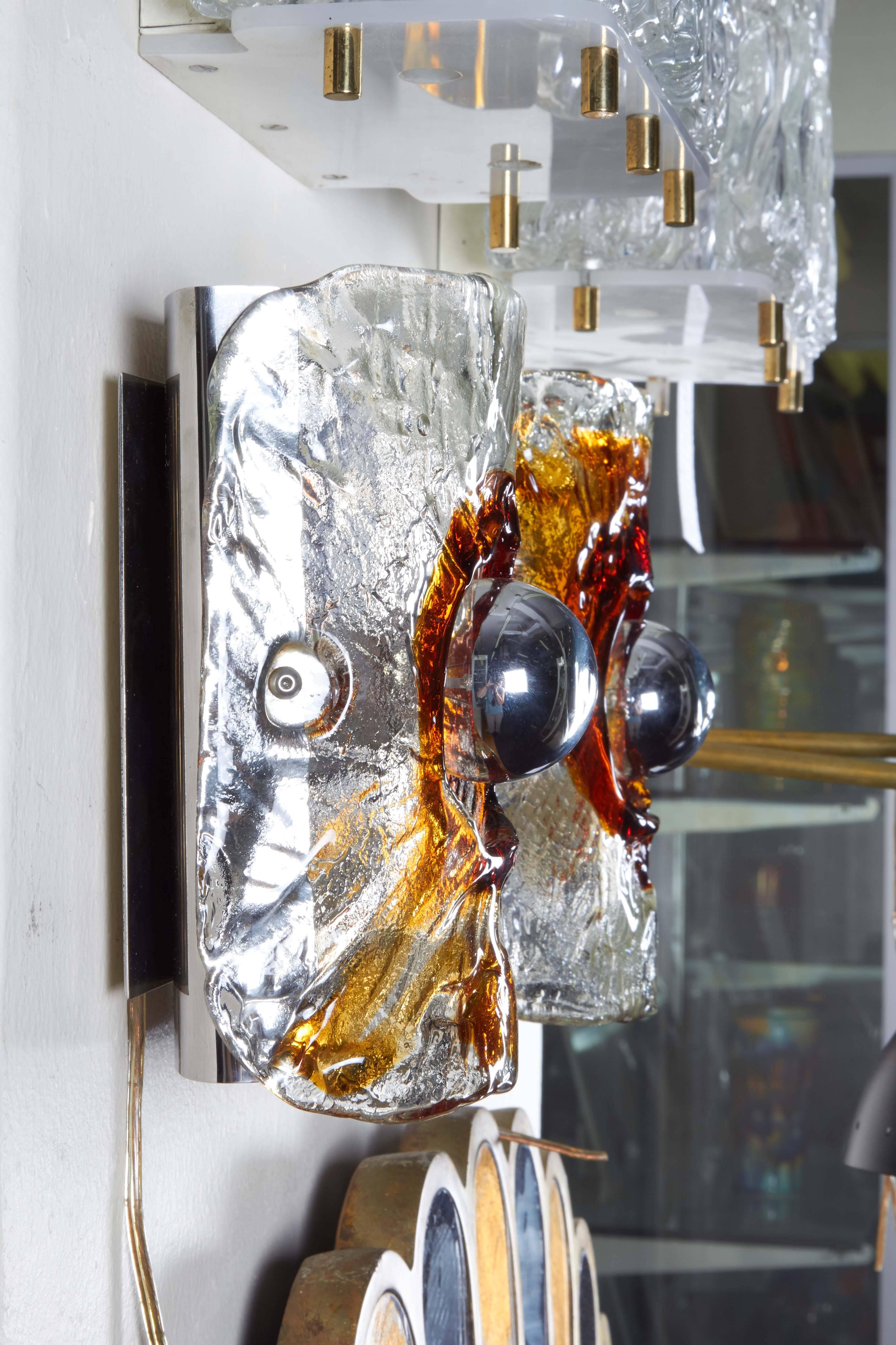 A pair of Italian wall sconces by Mazzega, produced, circa 1960s, each with convex shades in textured clear and amber Murano glass, surrounding a single exposed socket, affixed to a chrome backplate. Wiring and sockets to US standard. These light