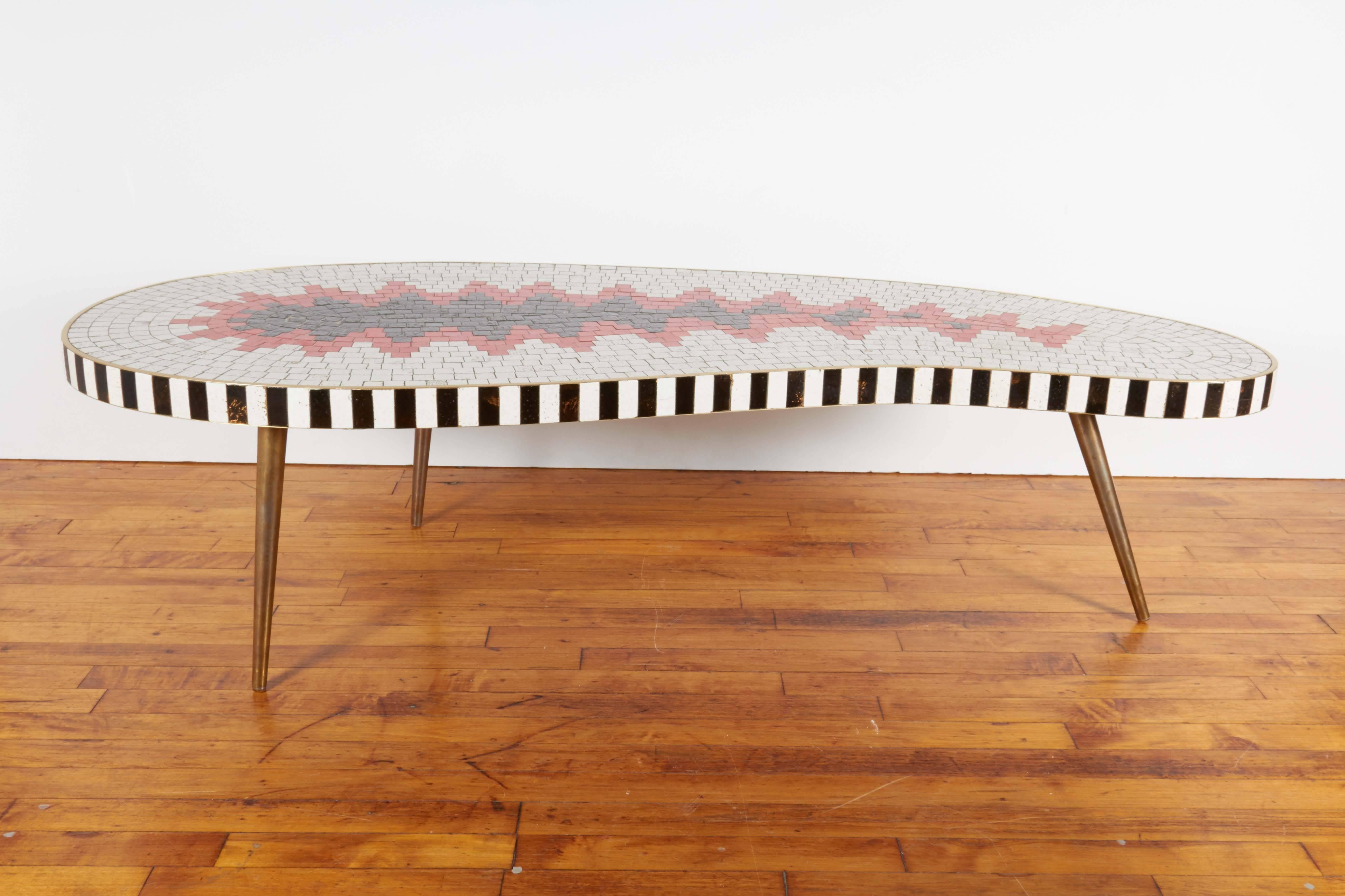 A kidney form coffee table, circa 1960s with mosaic top, composed of glazed ceramic tiles, in white, black and red, bordered by black and white pattern, set within a brass frame, on three tapered steel legs. The table remains in good vintage