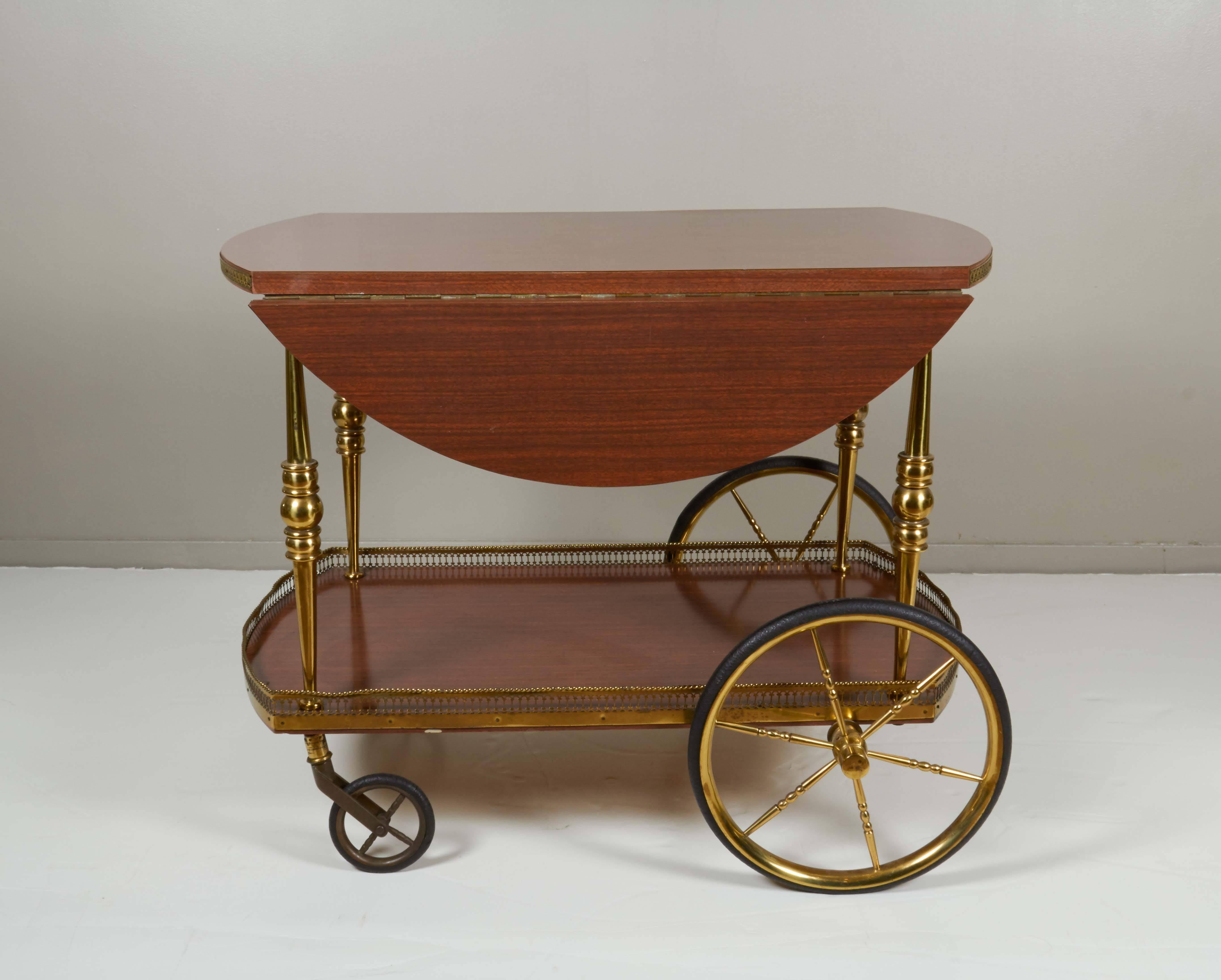 This Italian bar cart, produced circa 1960s, includes two-tiers with laminate wood surfaces, the top with guilloche border and two drop leaves, the bottom with a gallery, on caster and spoke wheels. The overall good vintage condition, with age