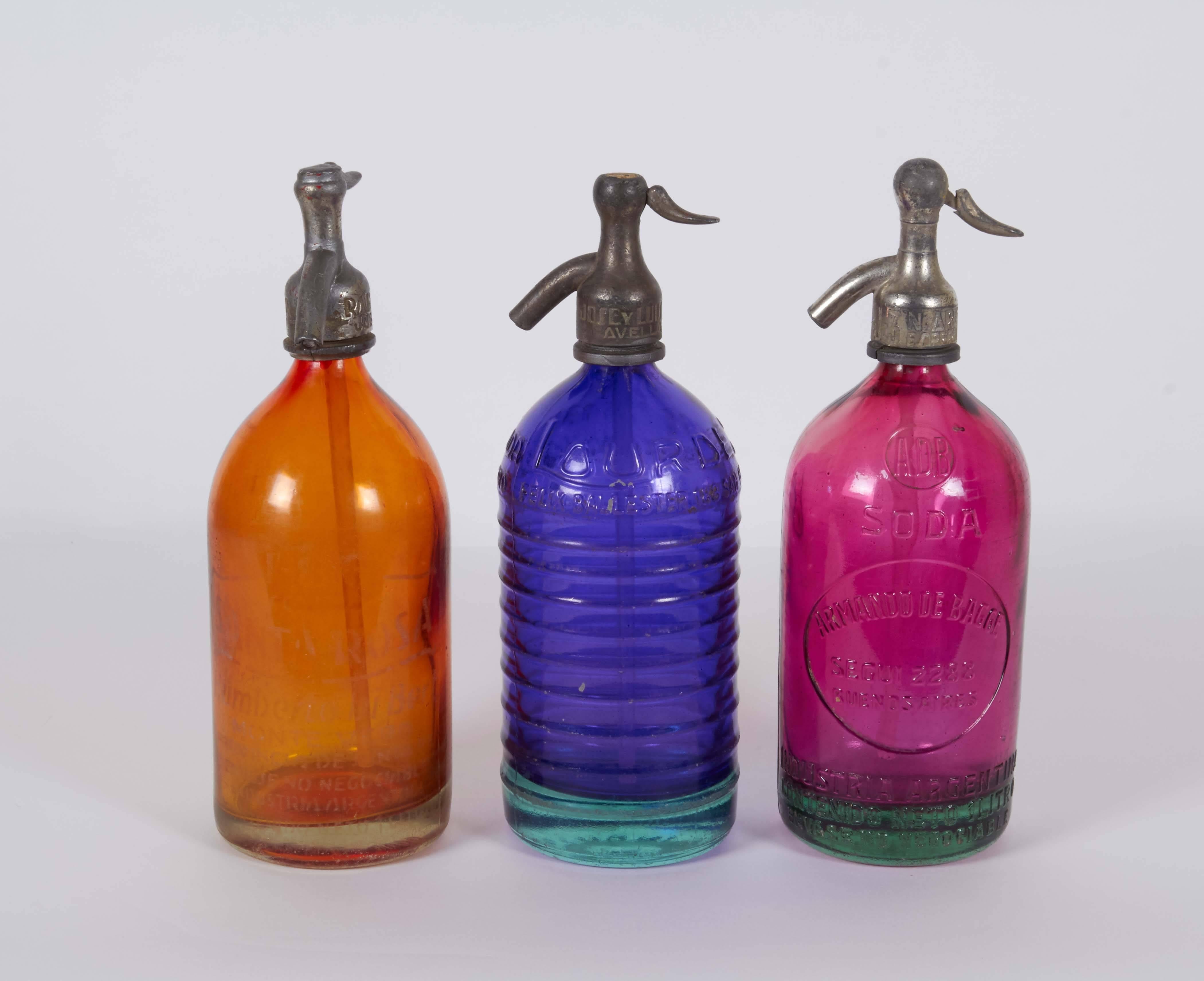 A set of three Art Deco era seltzer bottles, produced in Argentina, circa 1940s in vibrant colored glass, rare tones of orange, cobalt blue and fuchsia. Markings included to the metal nozzles and glass. The bottles remain in good vintage condition,