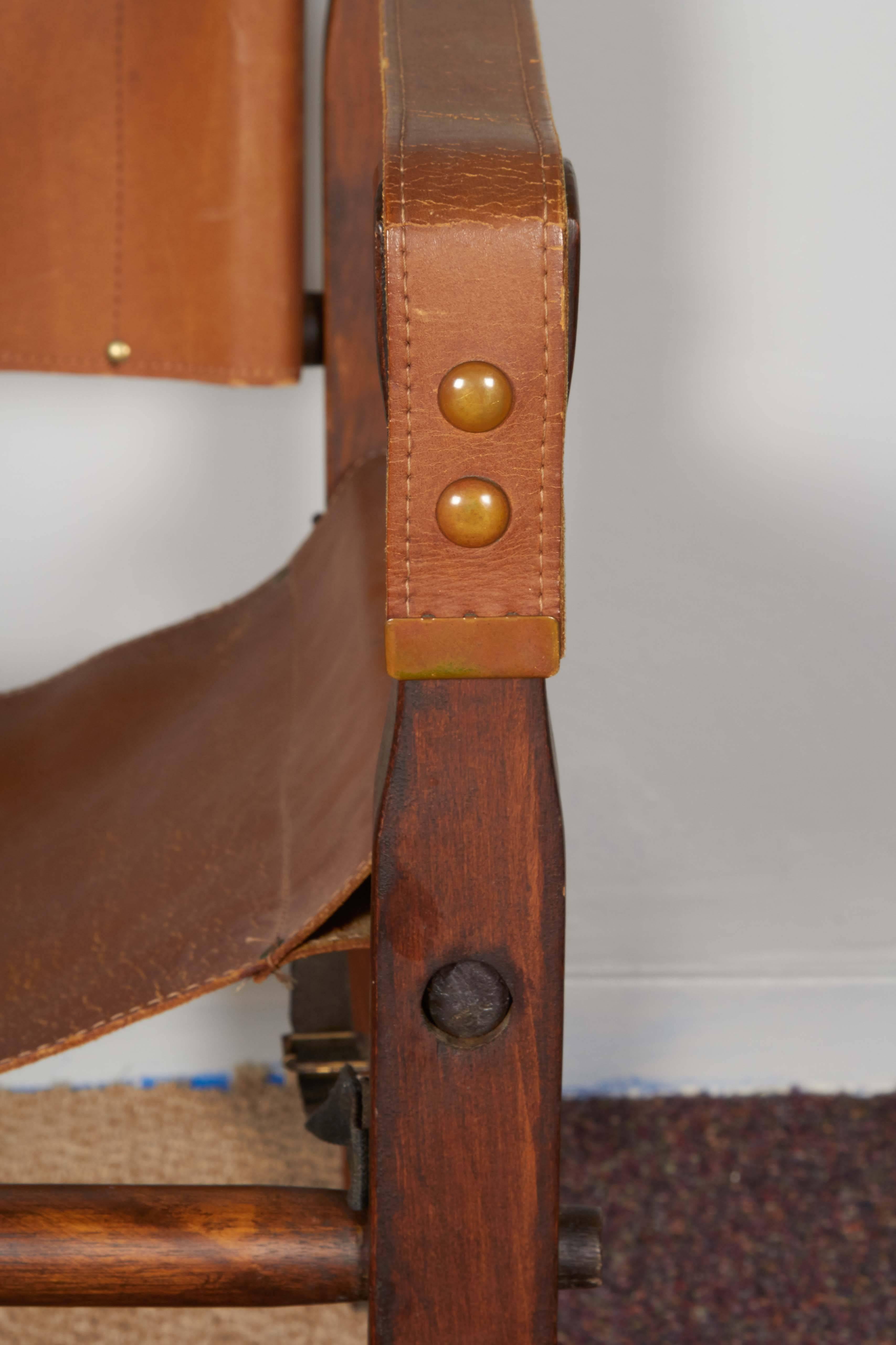 A pair of safari Campaign style chairs, produced, circa 1950s-1960s by the Gold Medal folding Furniture Company of Racine, Wisconsin, each with tan leather sling and arms, against warm toned wood frame. Markings include label to the back of one leg.