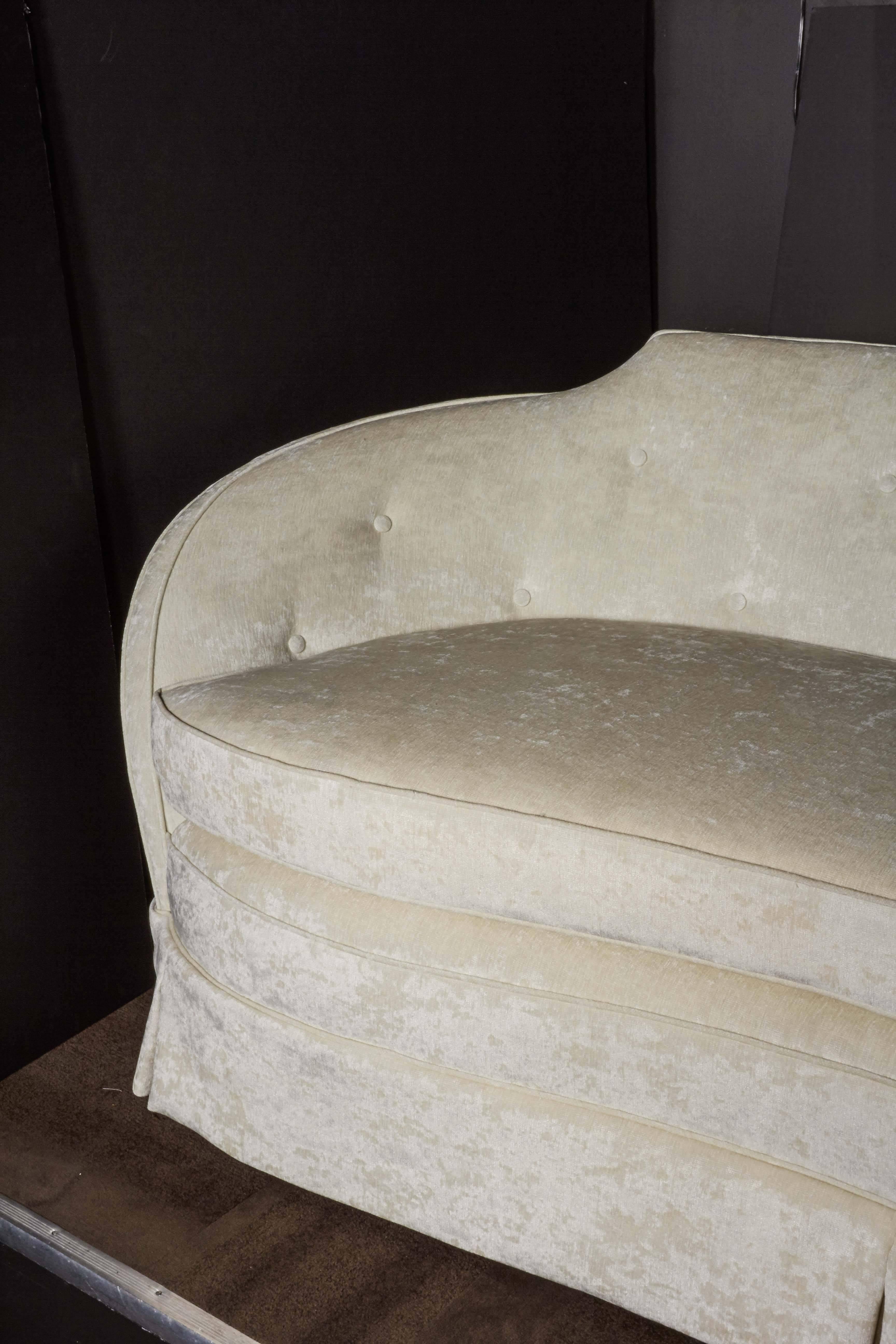 Striking Hollywood era sofa with stylized curved back/arm design. Newly reupholstered in a rich creme velvet by Castel, featuring button back details and dust ruffle. Comfortably sits up to four people.