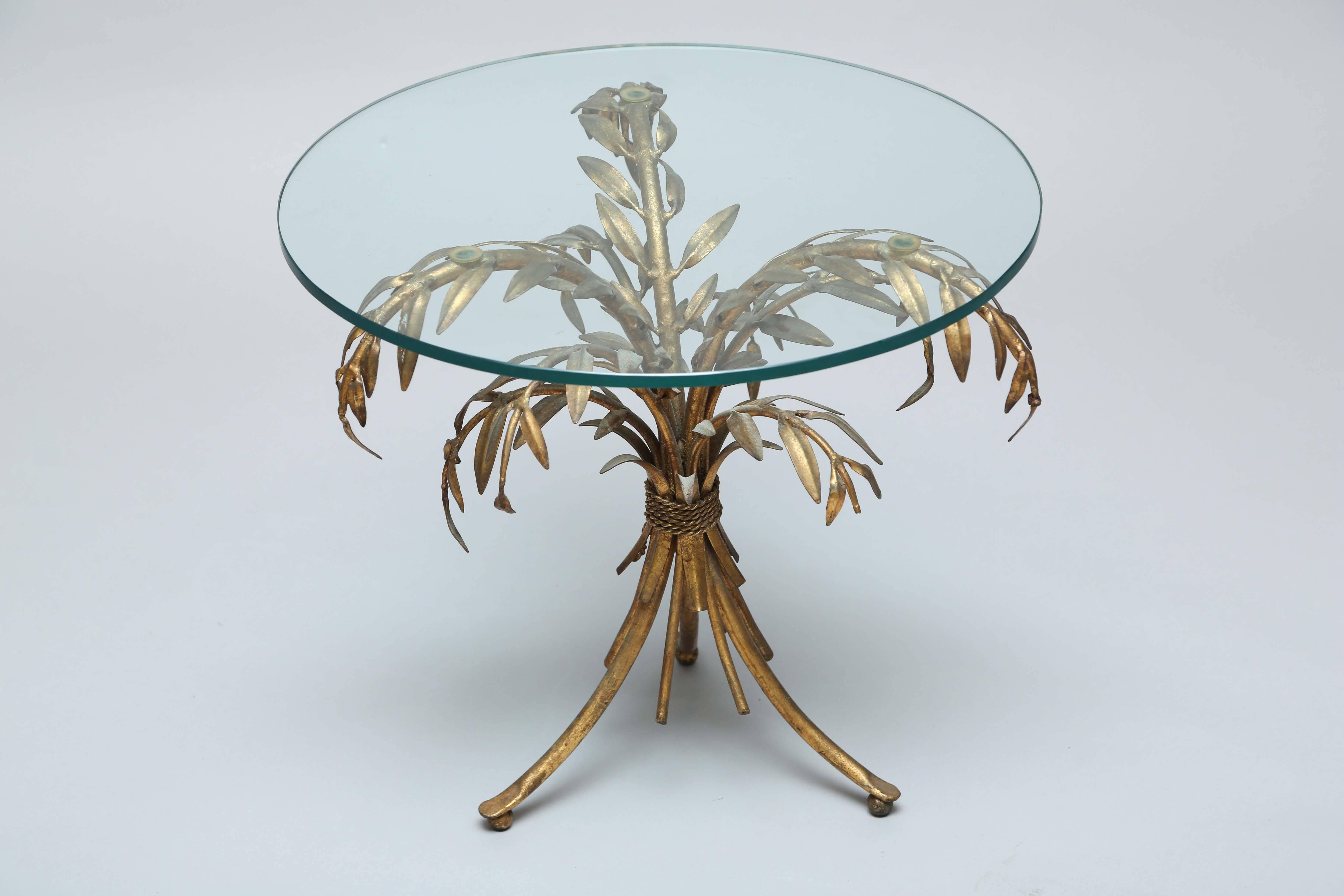 Accent table, having a round glass top, on gueridon base of gilded iron, shaped as palm leaves, tied at the center.