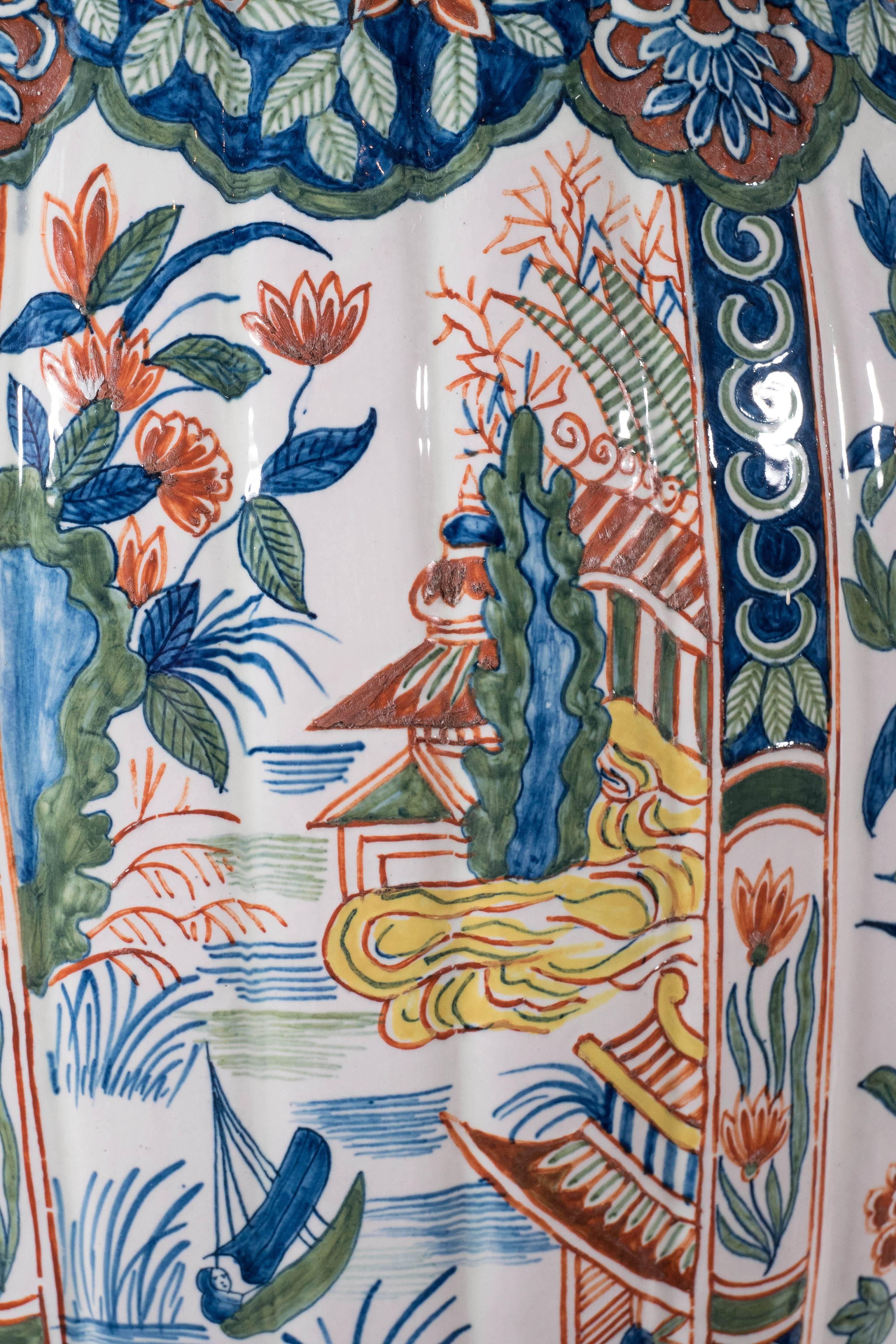 A pair of large polychrome jars with alternating panels. One panel shows a chinoiserie river scene with pavillions by the water. The other panel shows flowering branches in a garden. The vases are painted in a palette of greens, blues, orange, and
