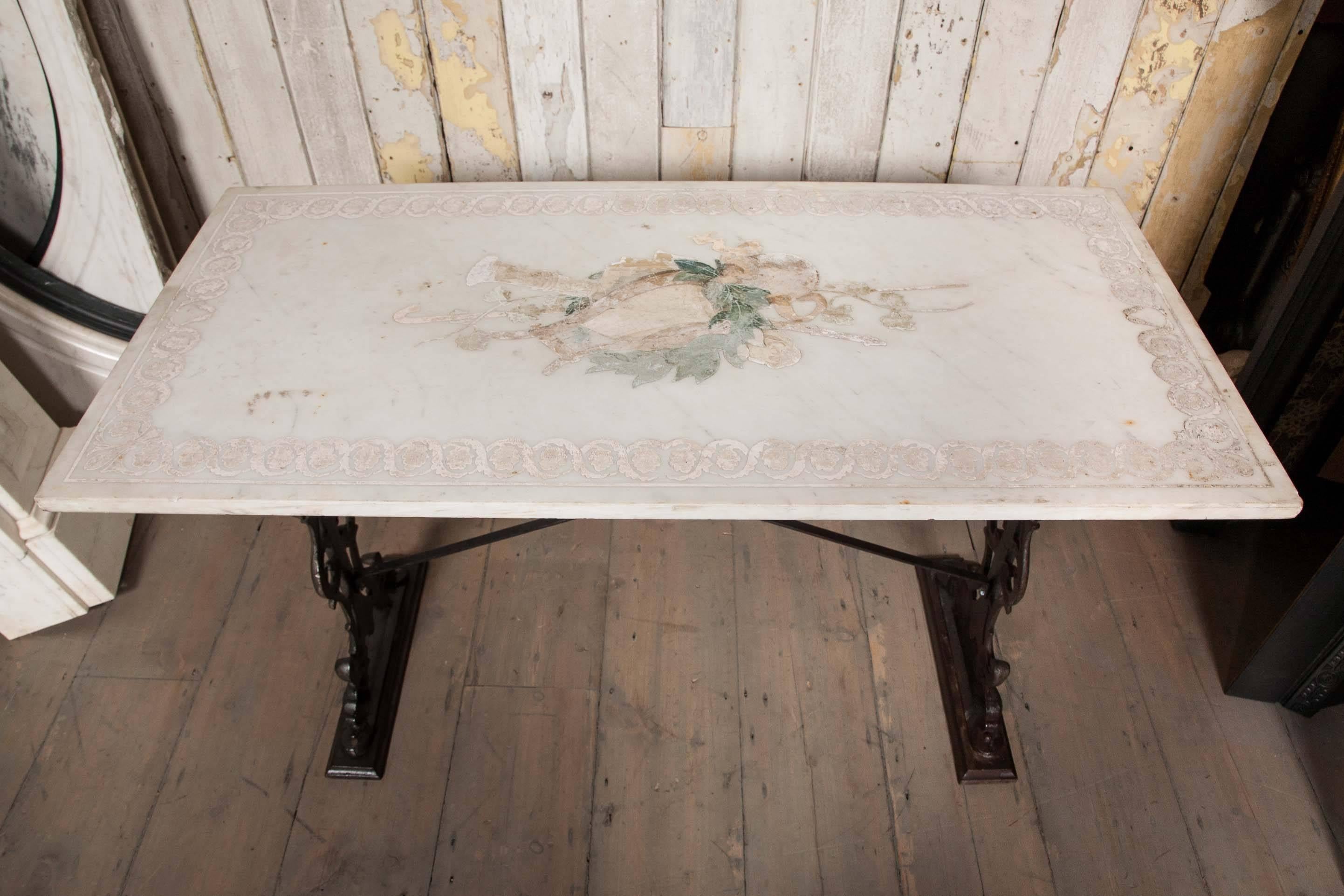 An original early Victorian marble tabletop on an ornately cast iron base. The table features a delicate neoclassical inlaid design with a lyre, a horn, a shepherd's crook and a wreath, surrounding by a floral border. The iron base is in the typical