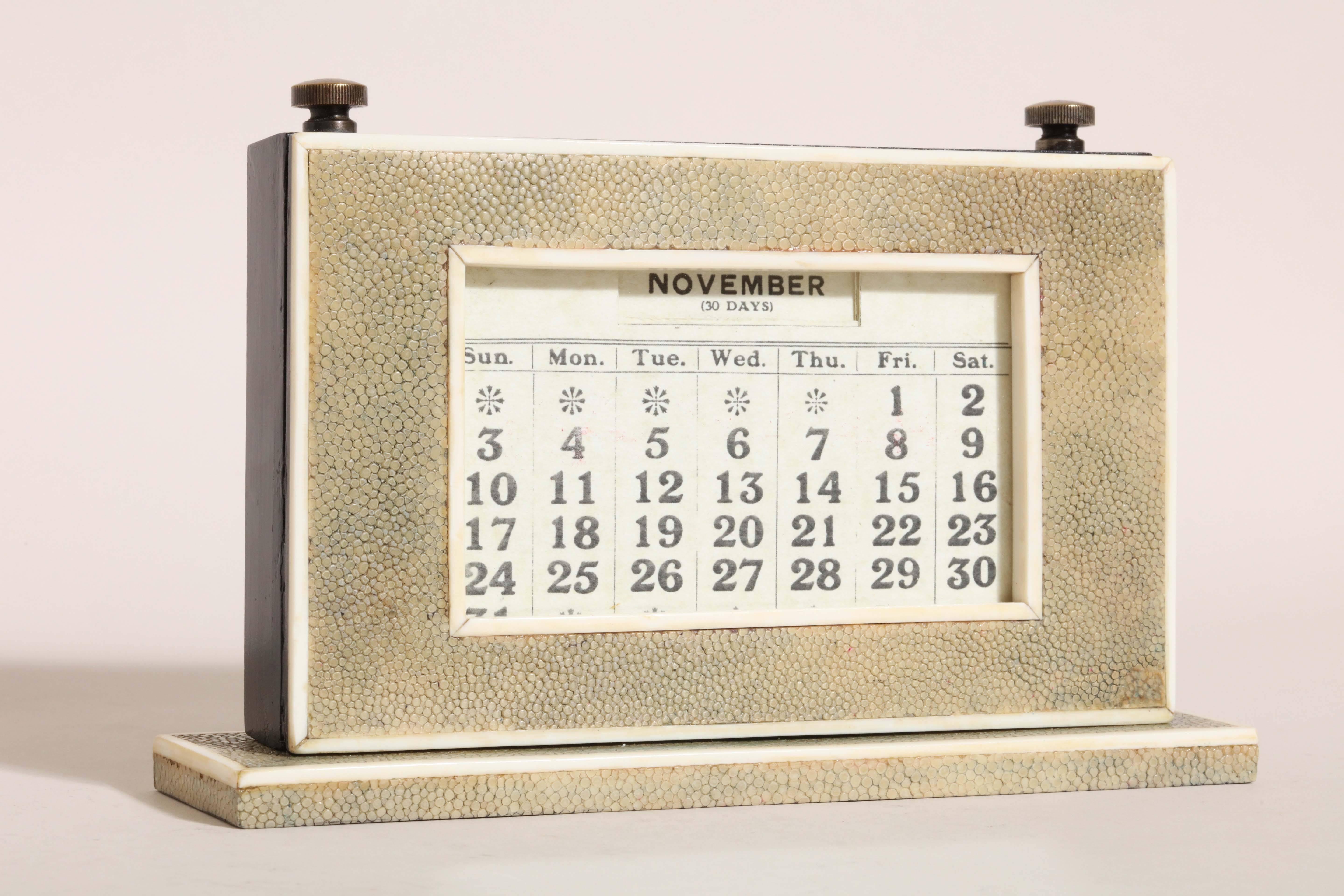 Wooden calendar covered in beige (with hints of blue) shagreen with bone banding around the base, the outside frame and the window. Has door in back to change the month cards. All cards are present.