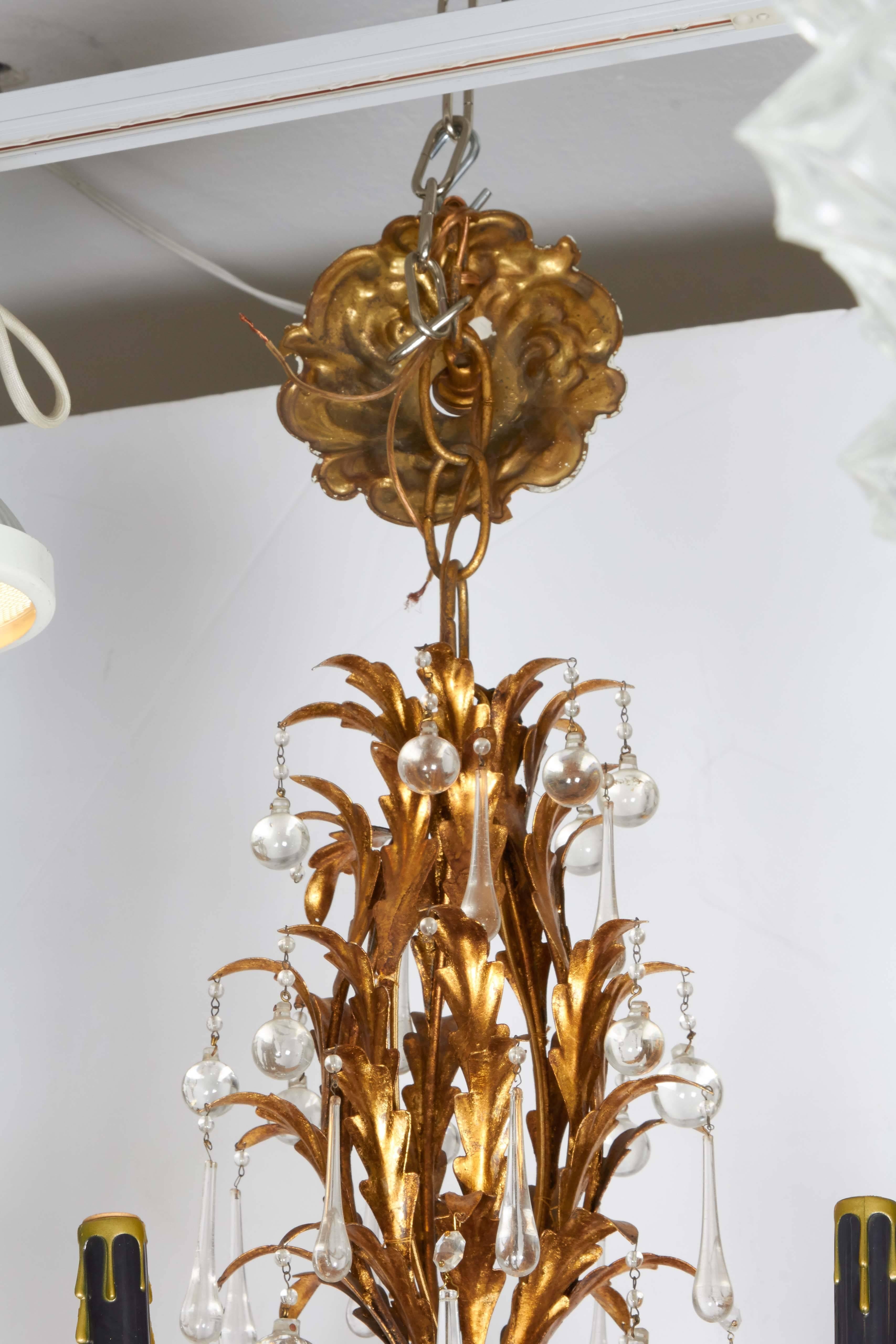 A luxurious tole chandelier in the Hollywood Regency style, with eight arms, each with black faux candle socket covers, on frame comprised of gilded metal foliage, trimmed with crystal drops. This light fixture remains in very good vintage