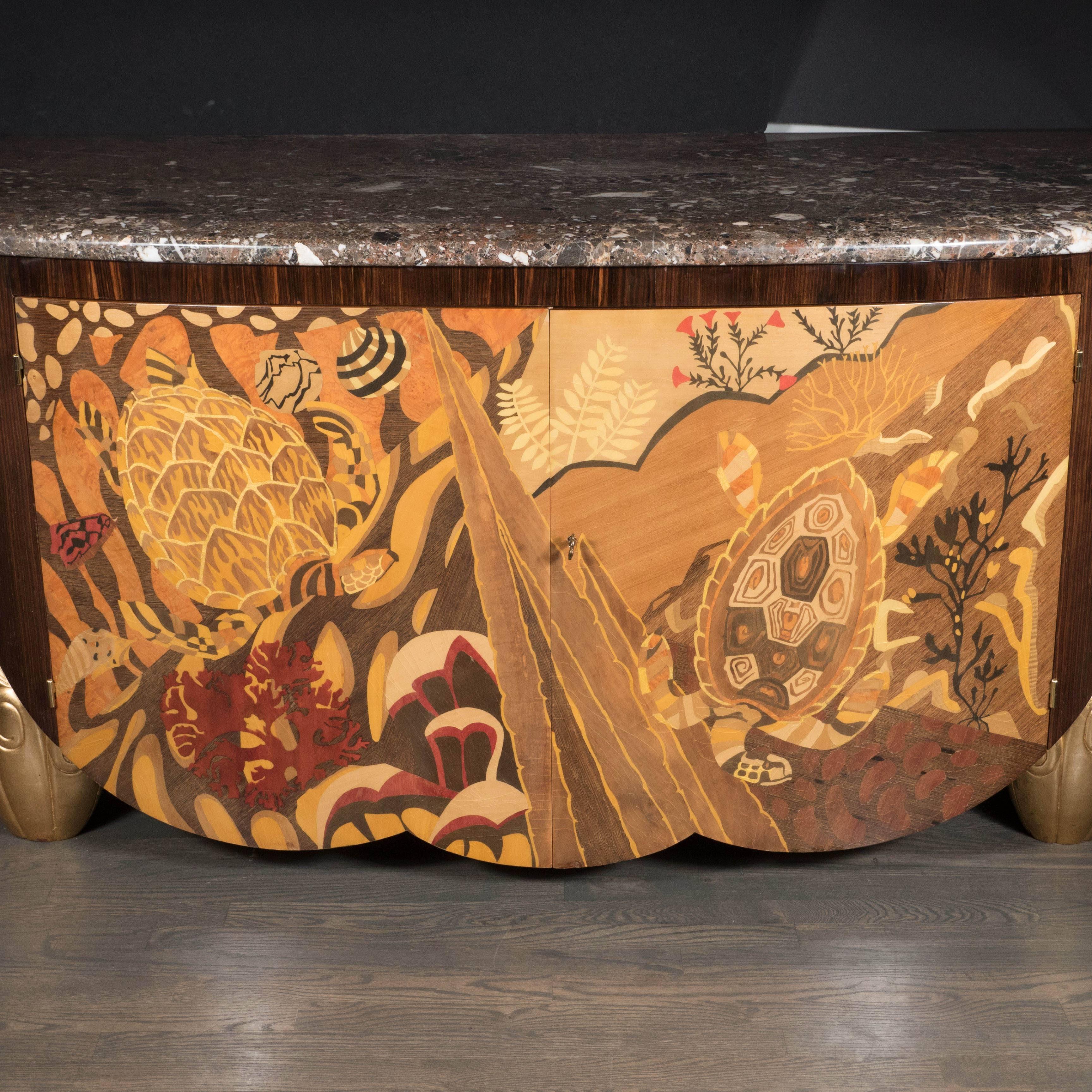 An Art Deco style sideboard or cabinet with exotic wood inlay and marble top. This demilune piece features two paneled doors which reveal three spacious pull-drawers. The facade features an intricate and detailed depiction of aquatic sea life
