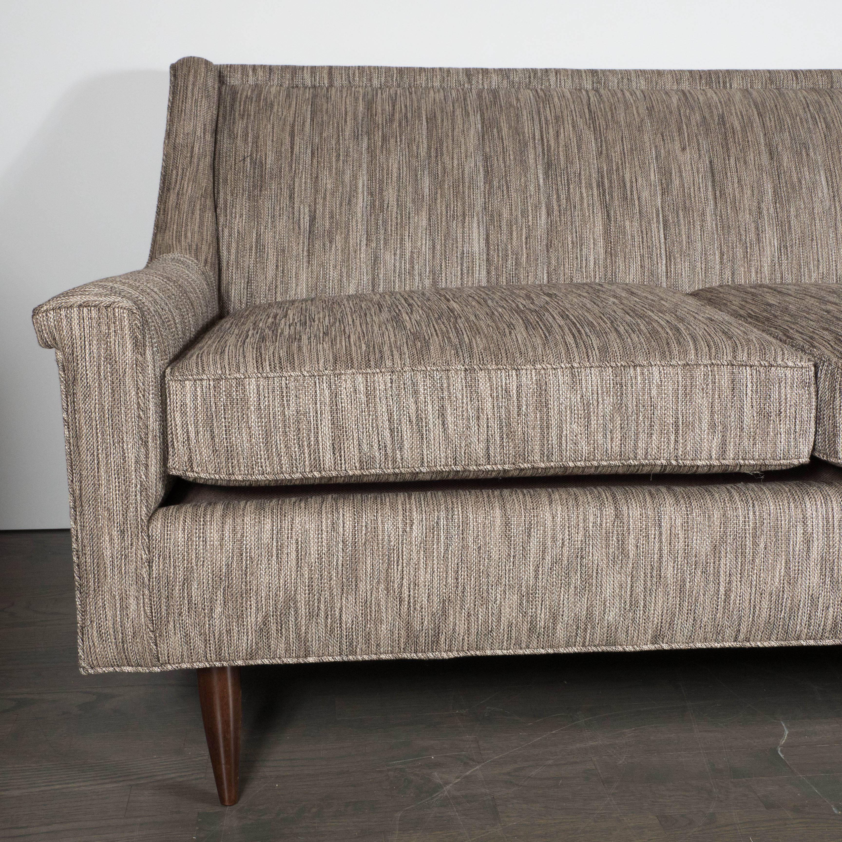 A Mid-Century loveseat in textured woven earth-toned upholstery in the manner of Dunbar. Conical walnut legs support a double removable cushioned loveseat. A vertically ribbed back is anchored by two sloped arms which fold outward, giving the piece