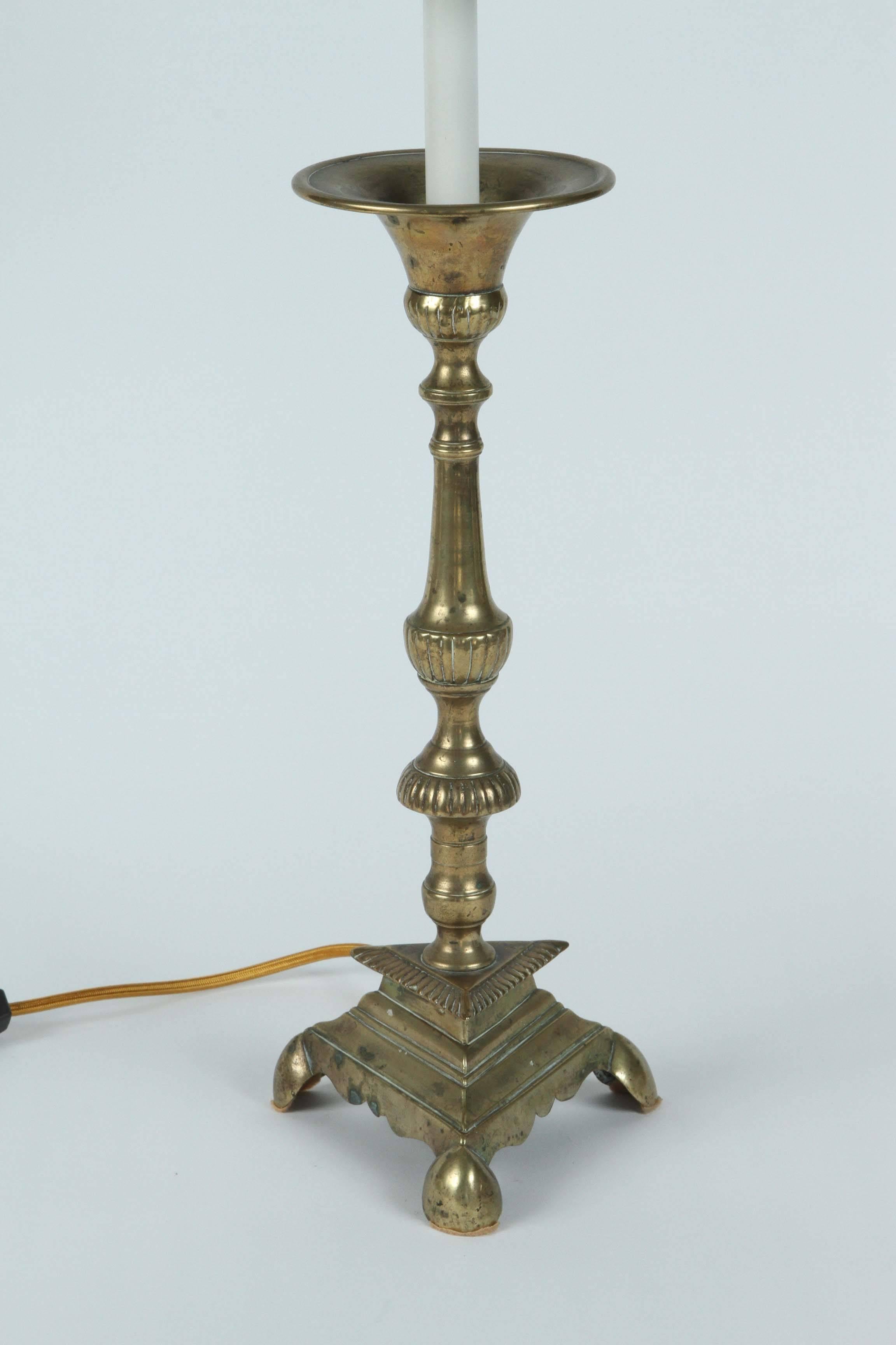 A single 19th century brass continental candlestick converted to lamp with custom linen shade. Details include an upswept drip pan and an unusual scalloped triangular base on oval feet.