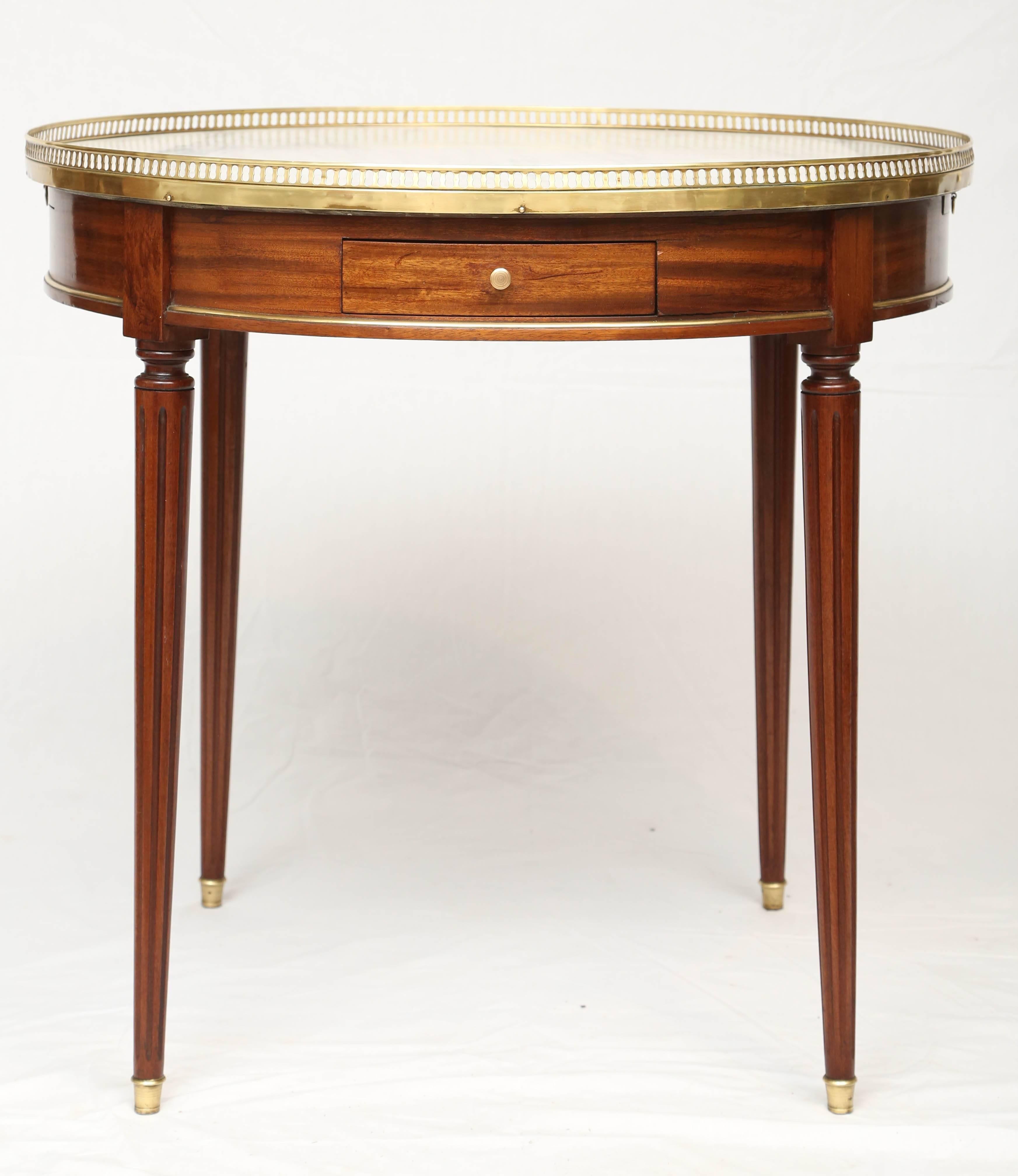 Bouillotte table, having a round top of white marble, surrounded by pierced gallery of polished brass, its apron revealing double slides, and two drawers, raised on round, tapering, fluted legs, ending in brass feet.