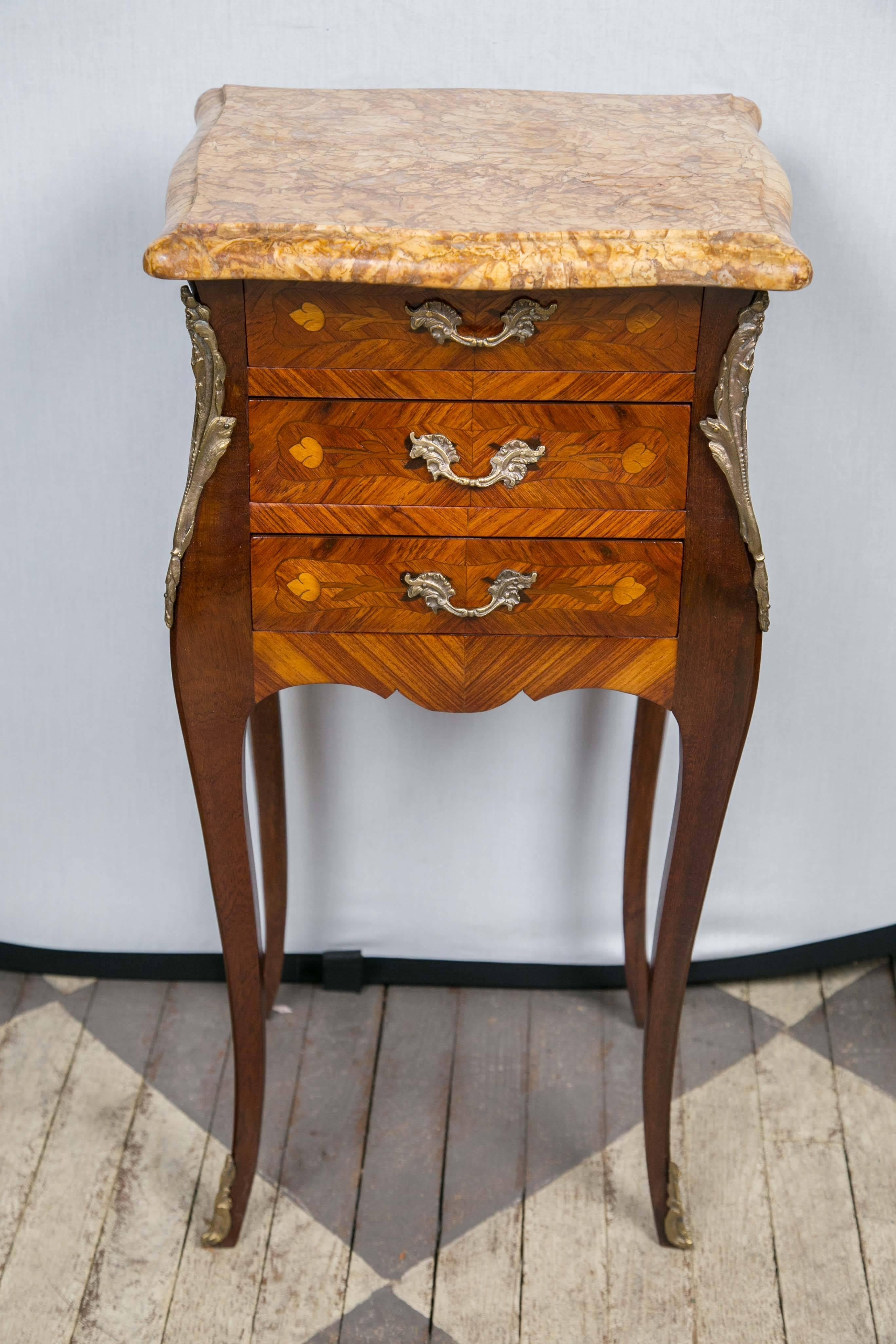 These are veneered in chevron pattern with inlays of simple flowers on the three-drawer fronts. Done in the Louis XV style, they have cabriole legs ending in applied bronze sabots on the front legs only. Shaped marble top, with ogee edge in hues of