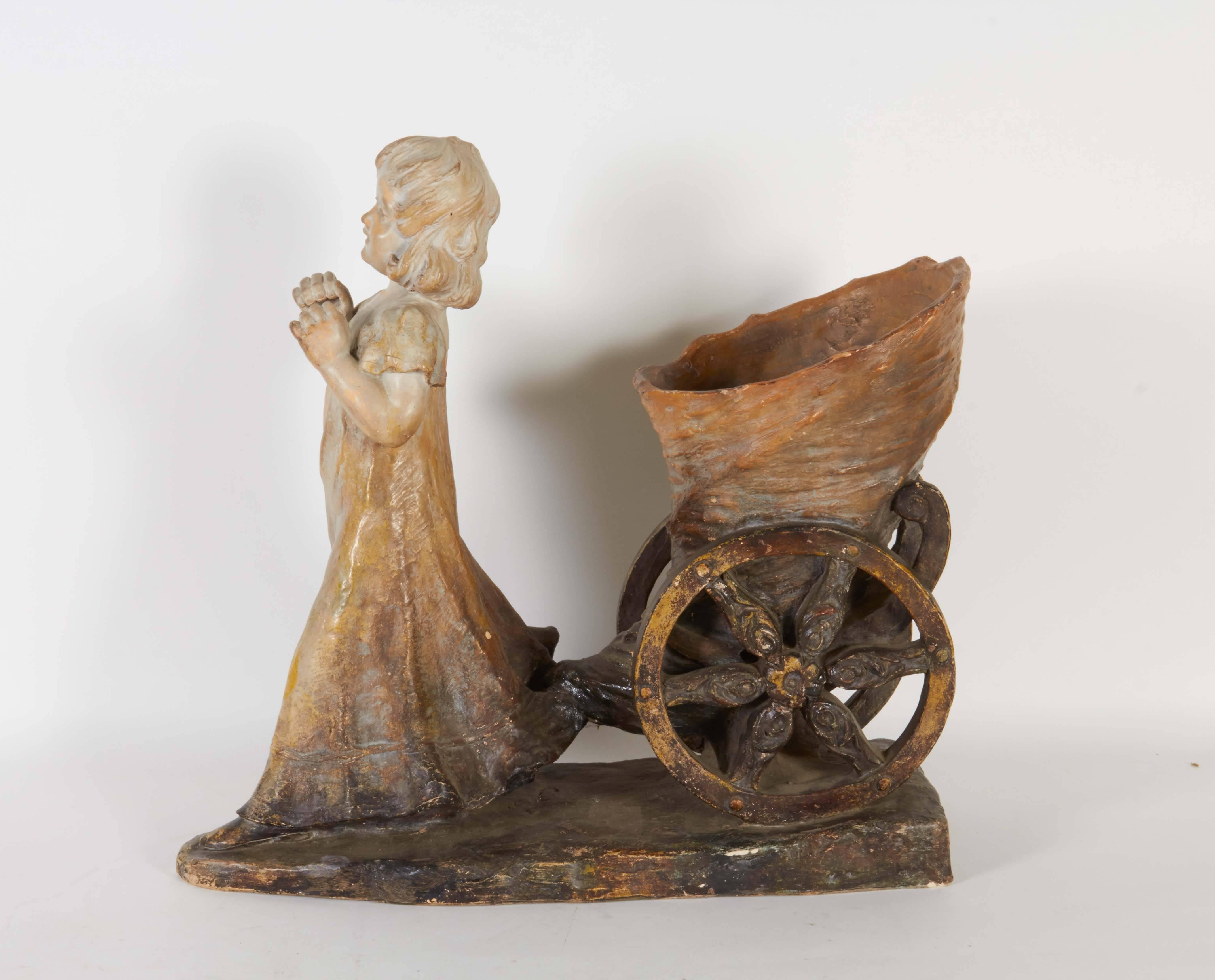 A beautiful terracotta jardiniere planter, produced in Austria within the early 1900s, depicting a sculpture of a young girl pulling a cornucopia on spoke wheels. Markings include [Made in Austria], number [300] and maker's name (albeit illegible)