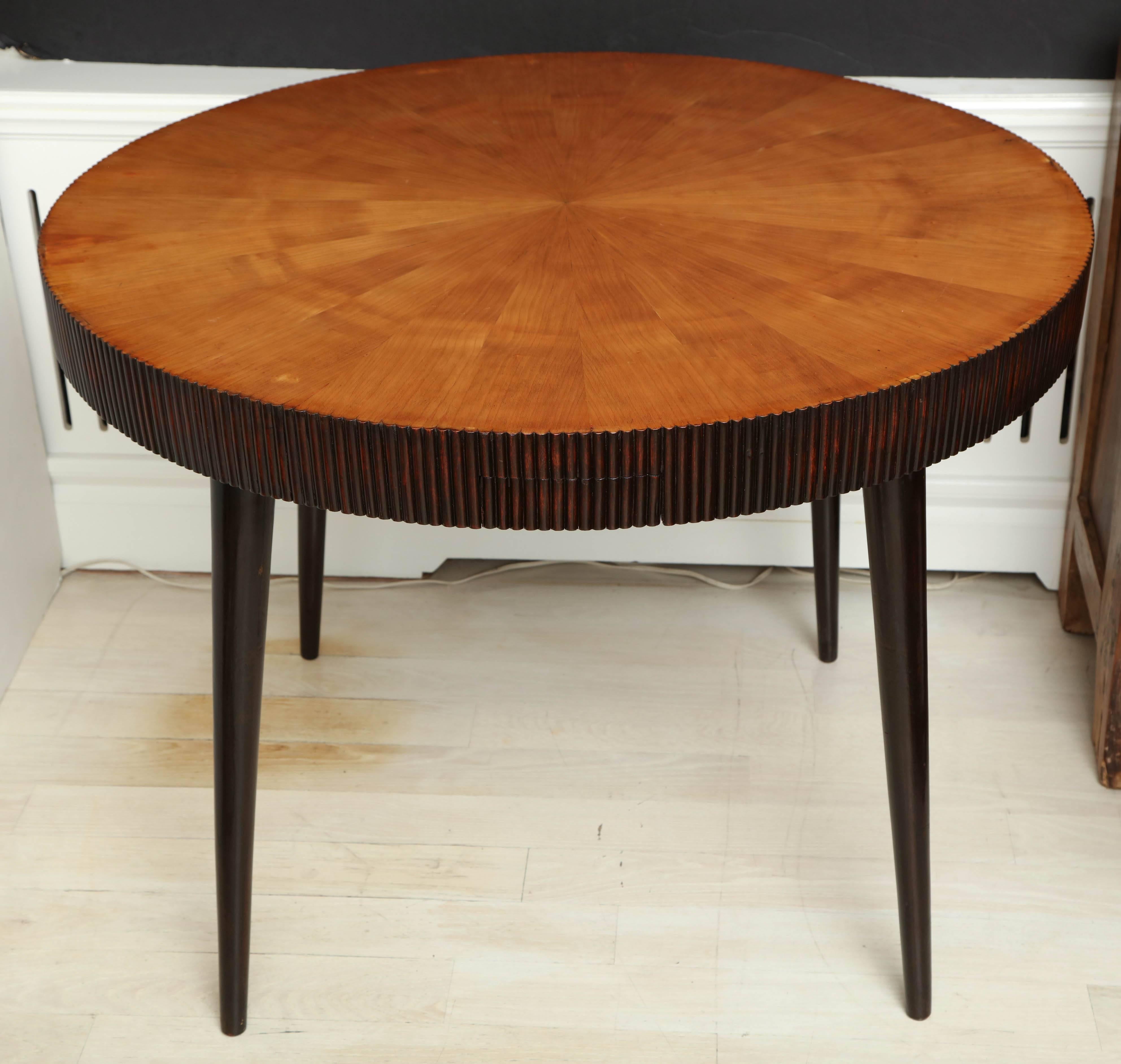 Round wood table with blond pie shape veneer, reeded edge and tapered legs, the reeded edge concealing four small drawers.

               