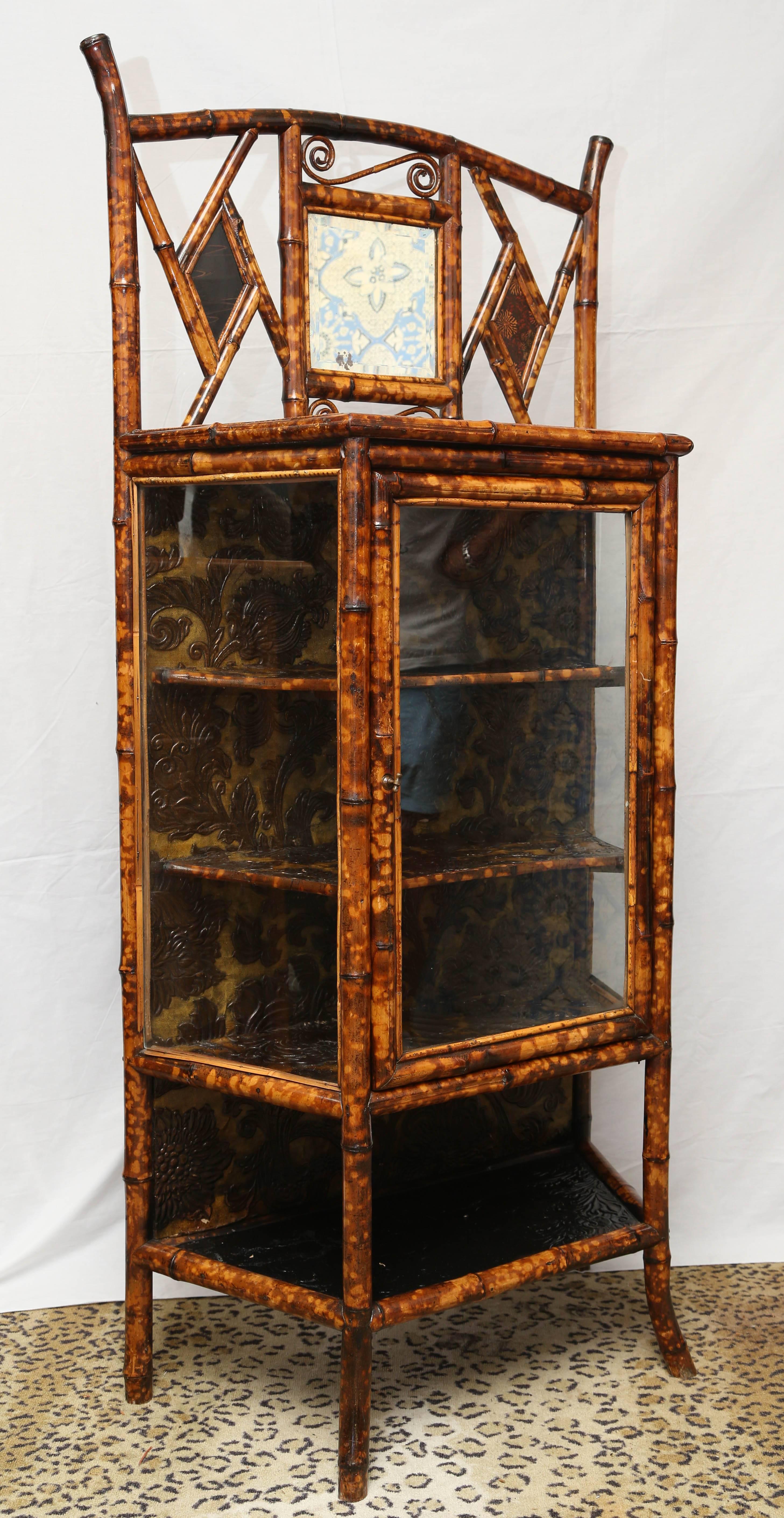 A nice English lacquer bamboo cabinet or bookcase, the pierced back inset with a beveled square mirror flanked by lacquered panels, the lacquered trapezoidal top with a glazed door opening to a shelved interior, over a medial shelf on Sabre legs.