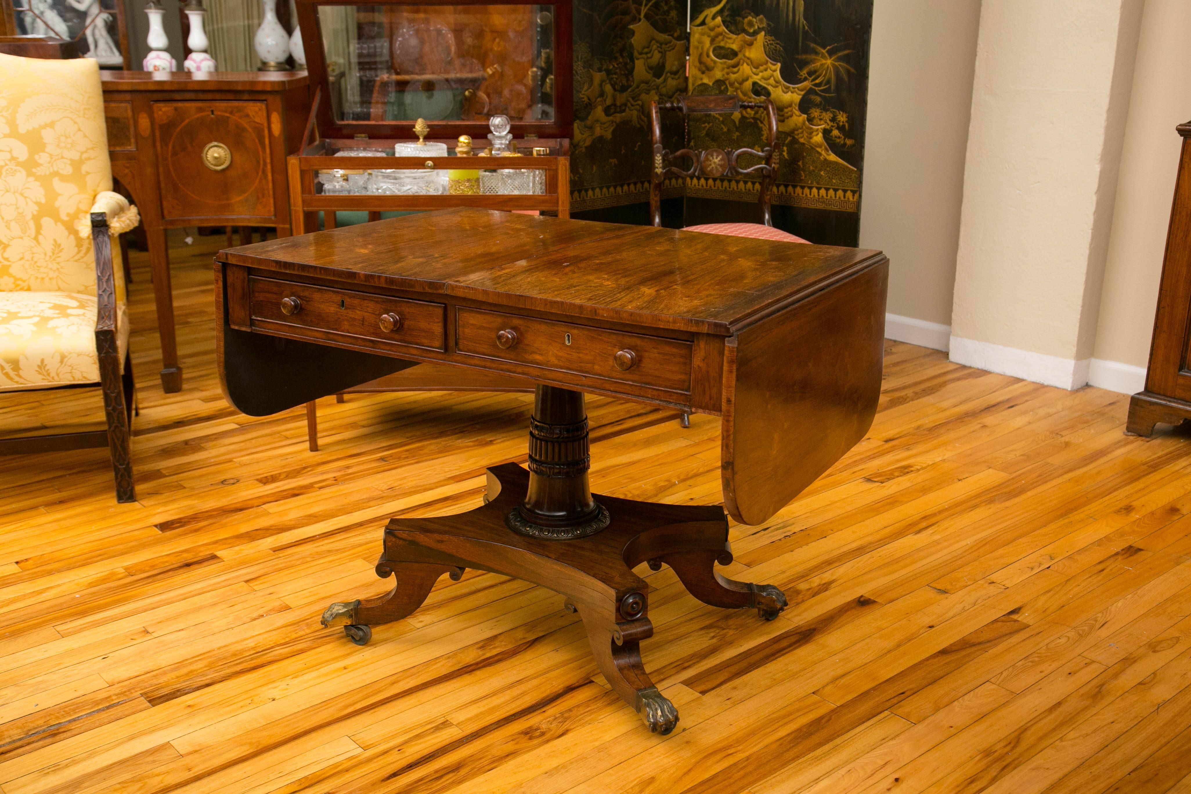 A fine English Regency sofa table over a quad-form base and four hipped splay legs capped with brass lion paw castors. Fitted with two drawers and having substantial drop down sides.