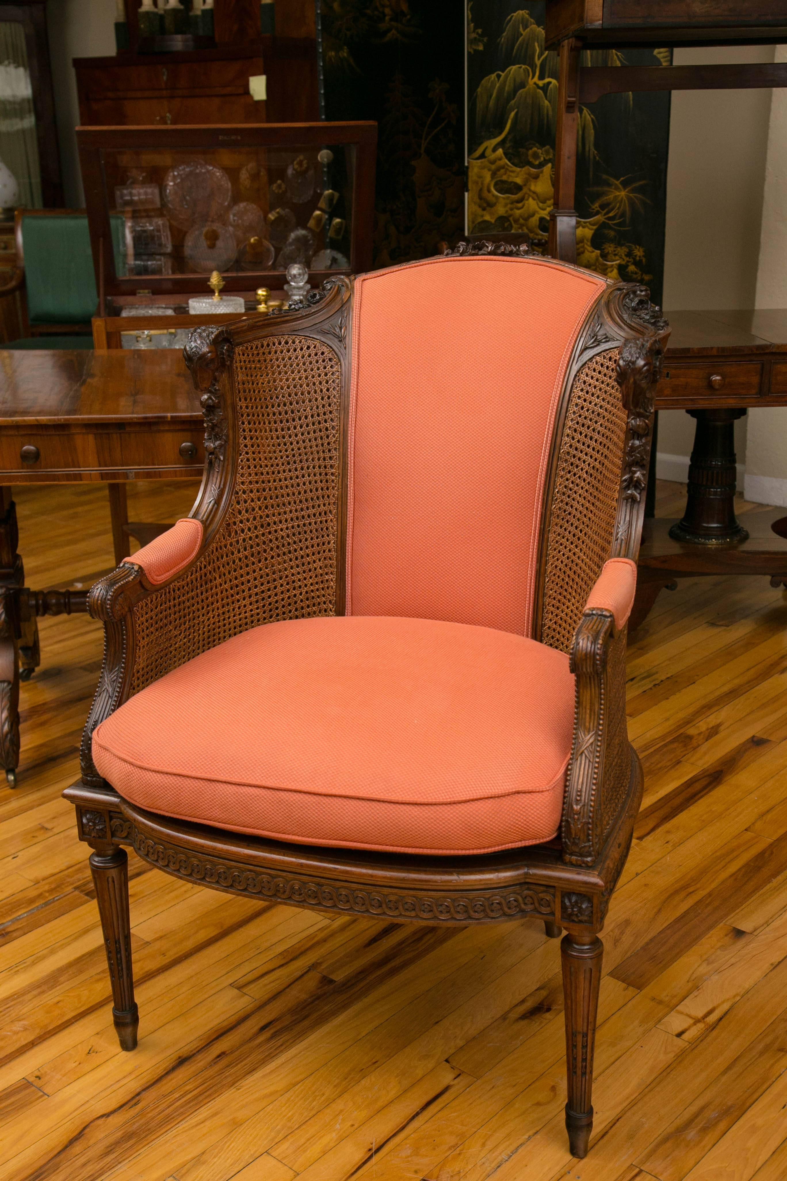 A distinguished hand-carved mahogany bergere library chair, with upholstered back and seat, caned sides with rams' heads adorning the crest rail.