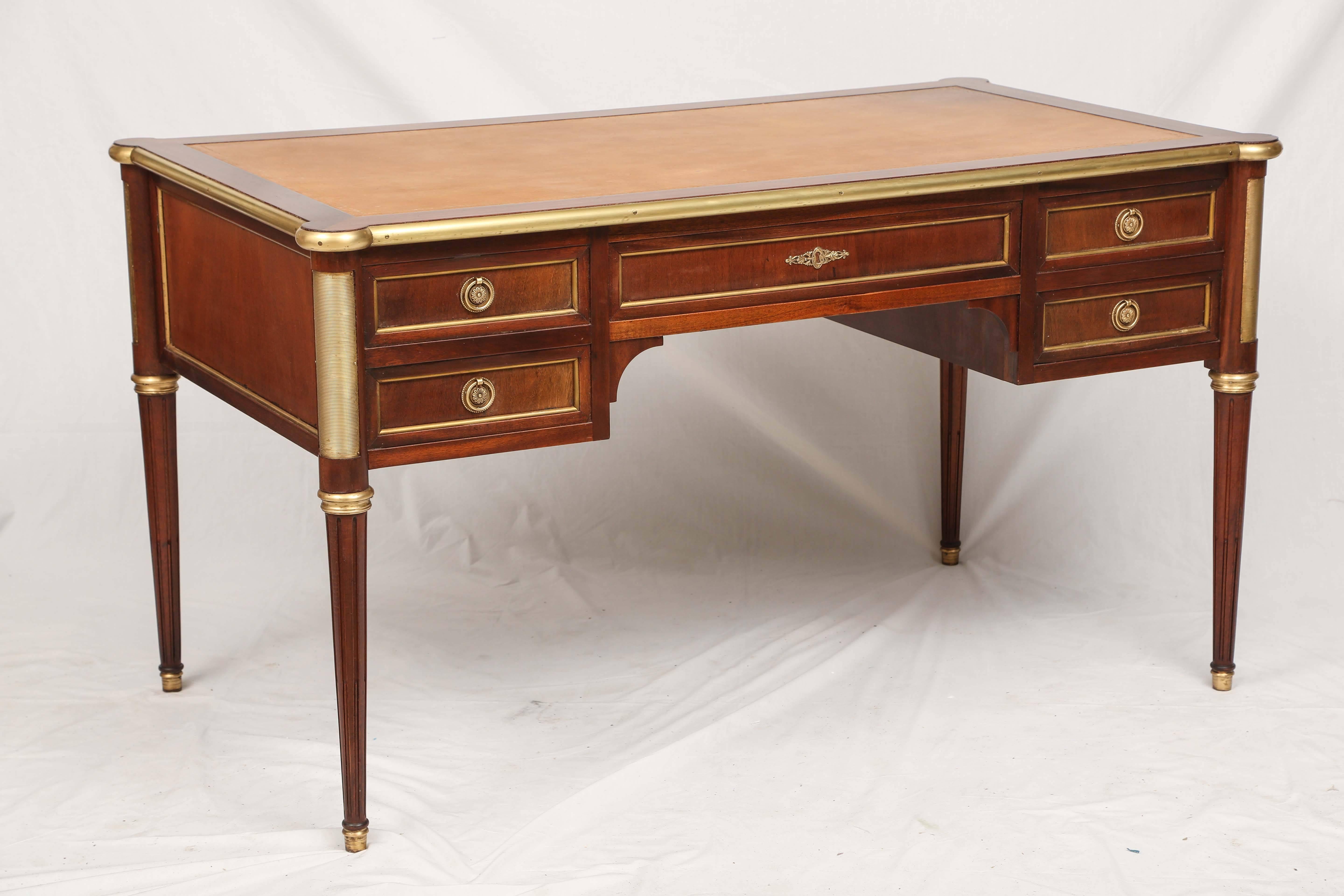 Classic Louis XVI style desk, of mahogany, having a rectangular inset leather top, bordered in brass, its case holds five fielded drawers, each with bronze pulls, rounded ormulated stiles at each corner, raised on tapering fluted legs, ending in cup